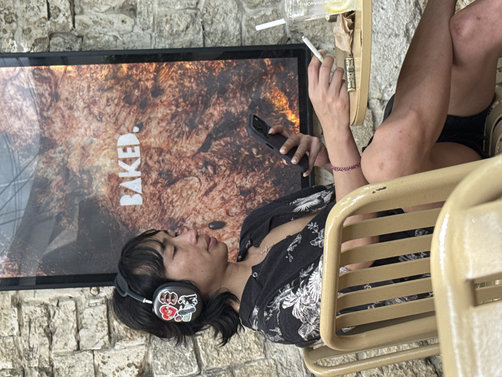 A young individual is captured in a relaxed moment, seated outdoors at what appears to be a cafe or restaurant in North Kuta, Indonesia. The person, with dark hair, wears large black over-ear headphones adorned with various stickers, including a red heart and a black cat-like figure. Their eyes are closed, suggesting they are engrossed in music or deep thought.
They are casually dressed in a dark button-up shirt with a light floral or bird pattern and black shorts. A pink and white braided wristband is visible on their left wrist. In their right hand, they hold a lit cigarette, and their left hand is gripping a black-cased smartphone.
The person is seated on a light-colored metal chair. On a wooden surface next to them, a clear plastic cup with the text "MORNING RITUALS" is partially visible, along with what looks like a lighter or accessory engraved with "FIRA."
In the background, a large framed poster or screen displays a textured, reddish-brown image with the word "BAKED." in white, block letters. Behind this, a light-colored stone wall provides the backdrop. The scene is well-lit, indicating it is daytime. The overall ambiance suggests a casual, outdoor setting where the individual is enjoying a break with music and a smoke.