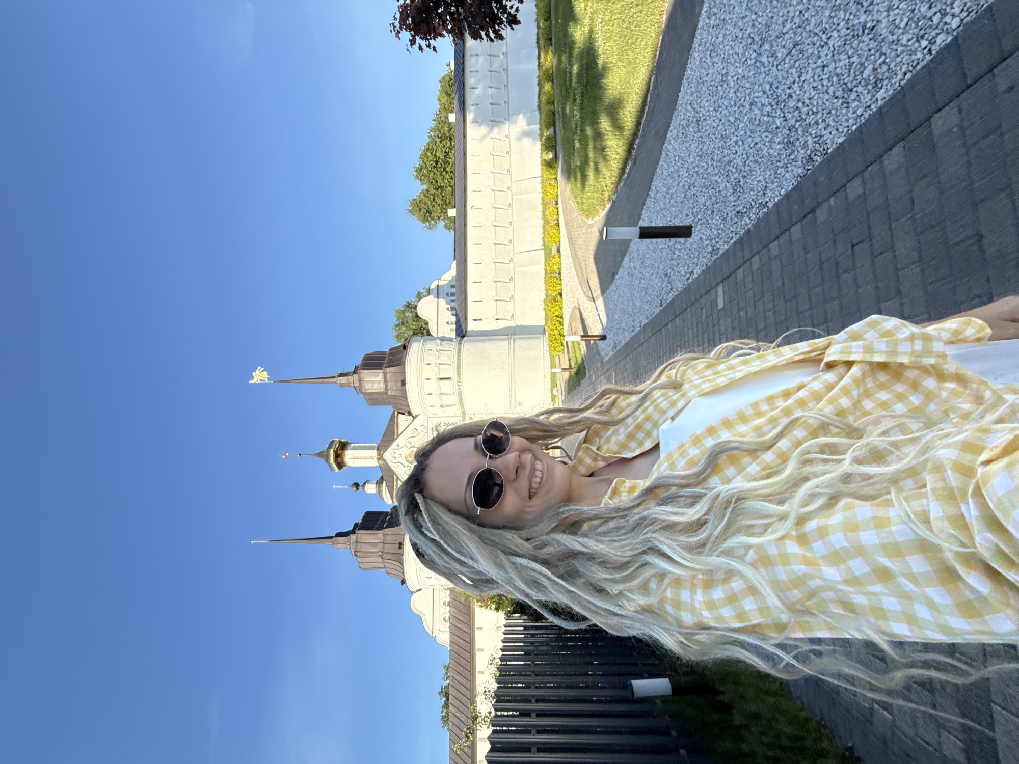 A smiling woman with long, light blonde dreadlocks and dark oval sunglasses is positioned in the foreground, facing slightly right and looking beyond the frame. She wears a yellow and white plaid button-up shirt over a white top. Behind her, a large white architectural complex, identified as being in the city of Dumino, Russia, features multiple towers with dark, conical spires and at least one prominent golden dome topped with a cross. A golden, stylized emblem adorns the peak of one spire on the rightmost tower. The complex is set against a clear blue sky. In the mid-ground, a paved pedestrian pathway with a light grey gravel border extends into the distance, flanked by neatly trimmed green grass and low green bushes. Several short, dark-colored pathway lights are spaced along the paved section. A dark metal fence is present on the left side of the frame. The scene is outdoors, under bright daylight.
