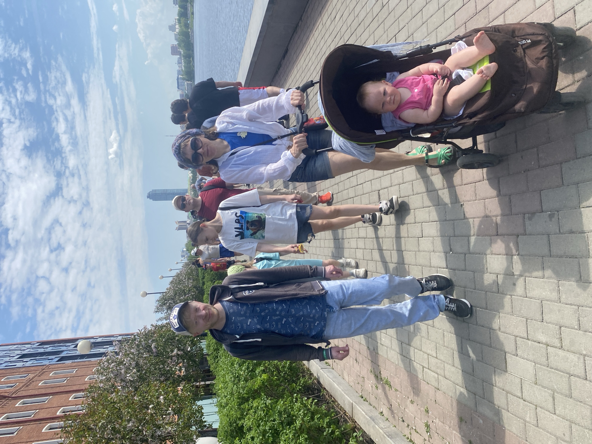 The image shows a group of people on a paved walkway next to a river. In the foreground, a man in jeans and a dark jacket is walking on the right side of the frame. Behind him, a young girl is walking, followed by a woman pushing a stroller with a baby inside. Other people are walking behind them, though less clearly visible. On the left side, there is a building and some greenery. In the background, the Iset River is visible. Across the river, there are buildings, including the Vysotsky skyscraper. Based on the presence of the Vysotsky skyscraper and the Iset River, this location is in Yekaterinburg, Russia. The weather appears sunny and bright.