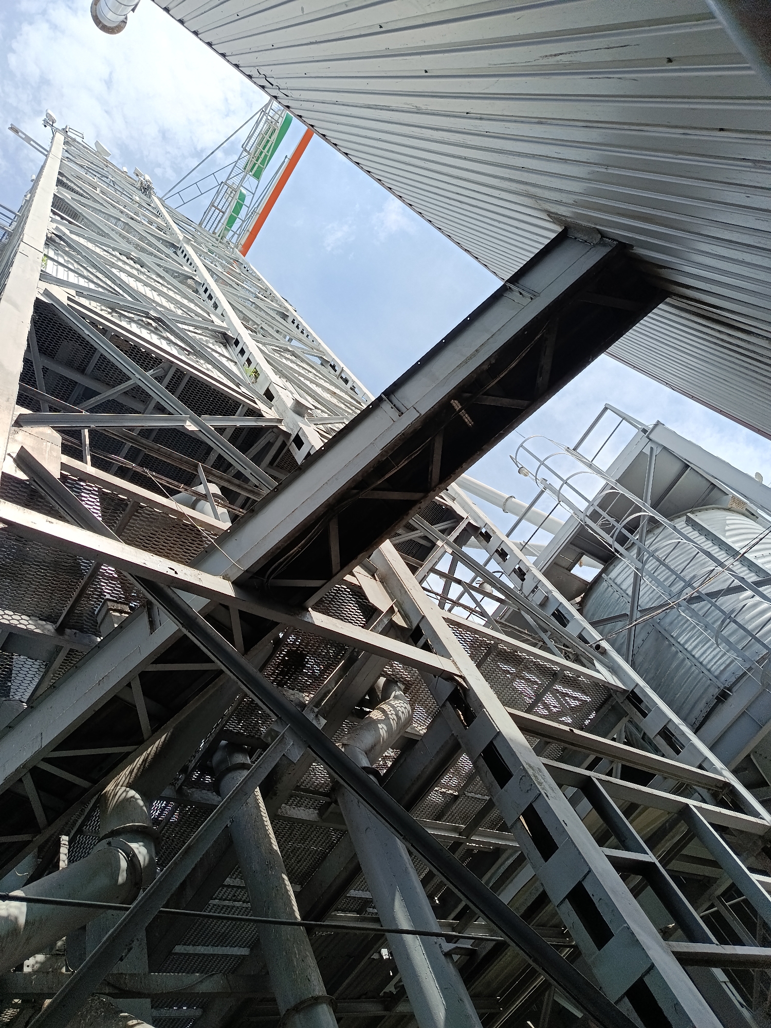 The image shows an upward view of a large industrial structure, likely part of a grain elevator, located in Biloziria, Ukraine. The structure is made of gray metal beams and grating, forming a complex network of platforms and supports. The sky is visible above, with some clouds. In the distance, a tall cylindrical silo can be seen. The perspective is from the ground, looking up towards the top of the structure. There are no people visible in the image. The overall scene is one of industrial architecture against a bright sky.