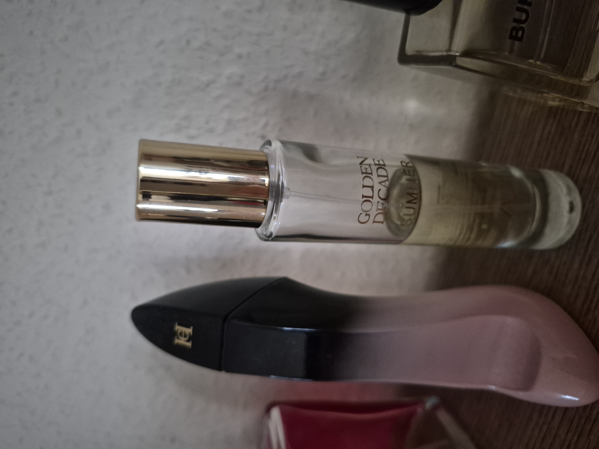 This is a close-up, eye-level shot of three perfume bottles on a wooden surface. From left to right, the first bottle is tall, black, and has a silhouette of a stiletto heel. The second bottle is shorter, clear glass with a gold cap, and contains a light yellow liquid. Text on this bottle reads "GOLDEN DECADE SUMMER". The third bottle is a square, clear glass bottle with a black cap, and the word "BUR" is visible on its front. The background is a textured, light-colored wall. The lighting is dim, and the focus is on the perfume bottles. The image appears to be taken indoors, on a shelf or dresser. There are no people or animals present.