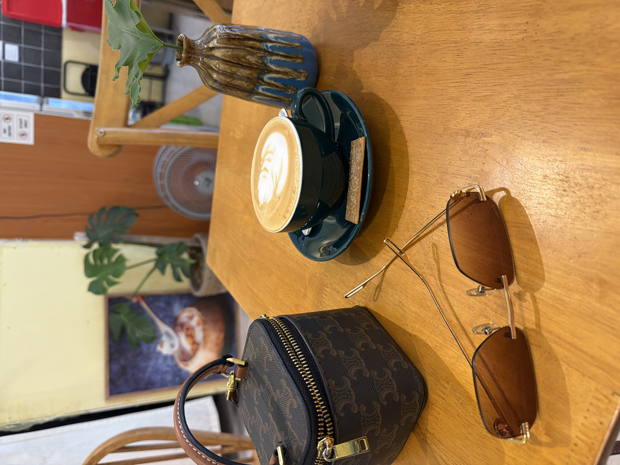 The image shows a tabletop scene in a likely cafe setting, possibly within Bac Nha Trang Ward, Vietnam, given the request. On the wooden table, there is a cup of coffee with latte art, placed on a green saucer with a small biscuit. Adjacent to the coffee are sunglasses and a small designer bag, possibly Celine. A decorative vase with greenery is also present. The background includes elements suggesting the location is an indoor seating area, like a fan, artwork, and other furniture, but they are slightly out of focus. The overall impression is a casual scene showcasing personal items alongside a beverage.