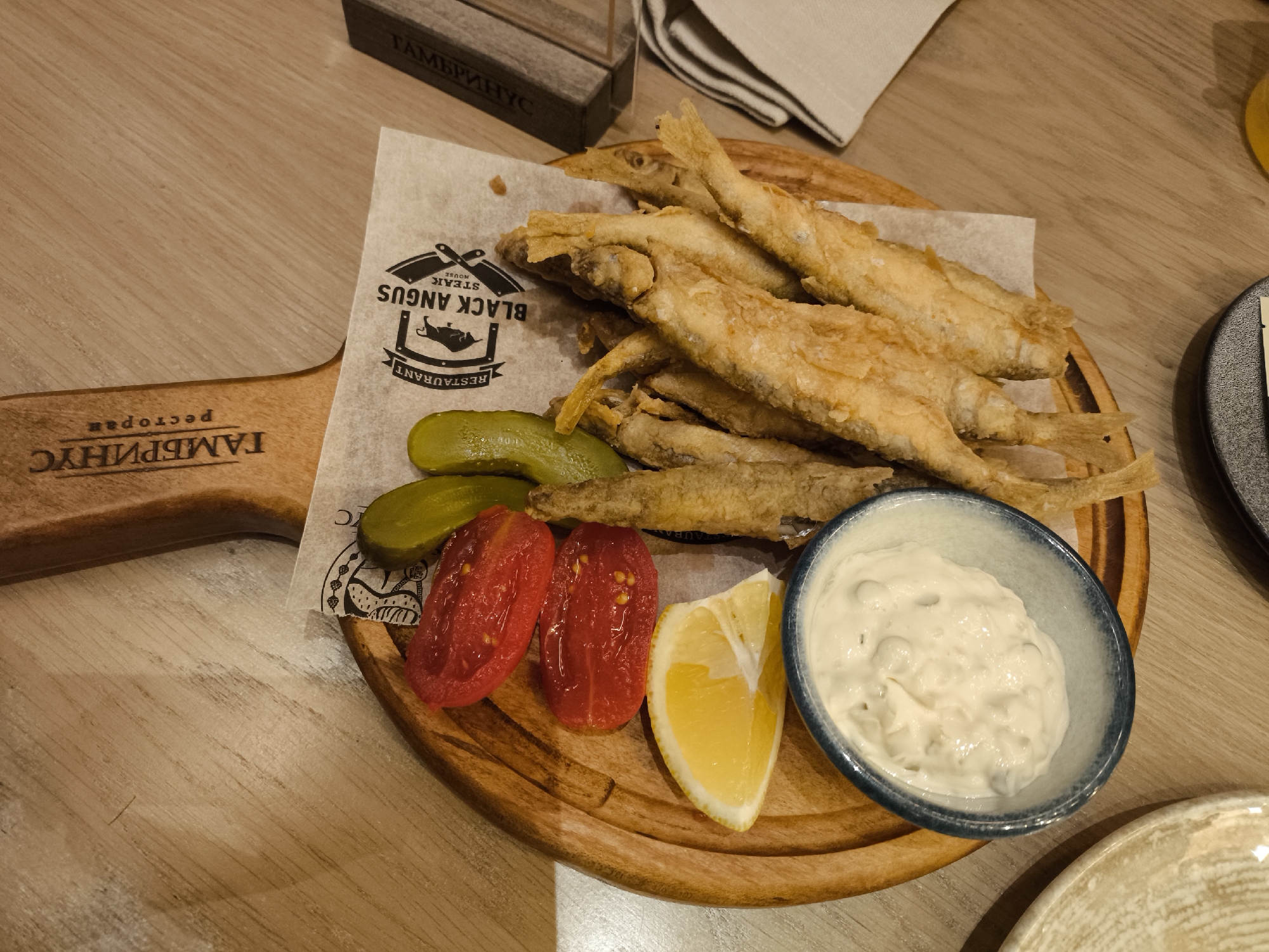 The image shows a wooden serving board with fried fish, pickled cucumbers, sliced tomatoes, a lemon wedge, and a small bowl of white sauce. The board also has a paper liner with the words "BLACK ANGUS STEAK HOUSE RESTAURANT". The handle of the board is visible, with Cyrillic text on it. The background shows a wooden table surface. Based on the "BLACK ANGUS STEAK HOUSE" logo and the Cyrillic text on the handle, it's likely that the location is Moscow, Russia.