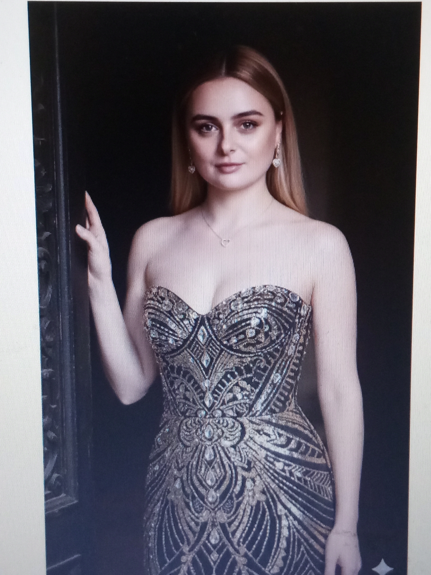 A young woman with blonde hair and fair skin, wearing a strapless, embellished gown and heart-shaped earrings, stands with her left hand resting on a dark, ornate doorway. Her right arm is bent, with her hand resting on her chest. She is looking towards the viewer with a subtle smile. The background is dark and indistinct. The image is a photograph, likely taken indoors. There is no information in the image to suggest the location is the city of Riga, Latvia.