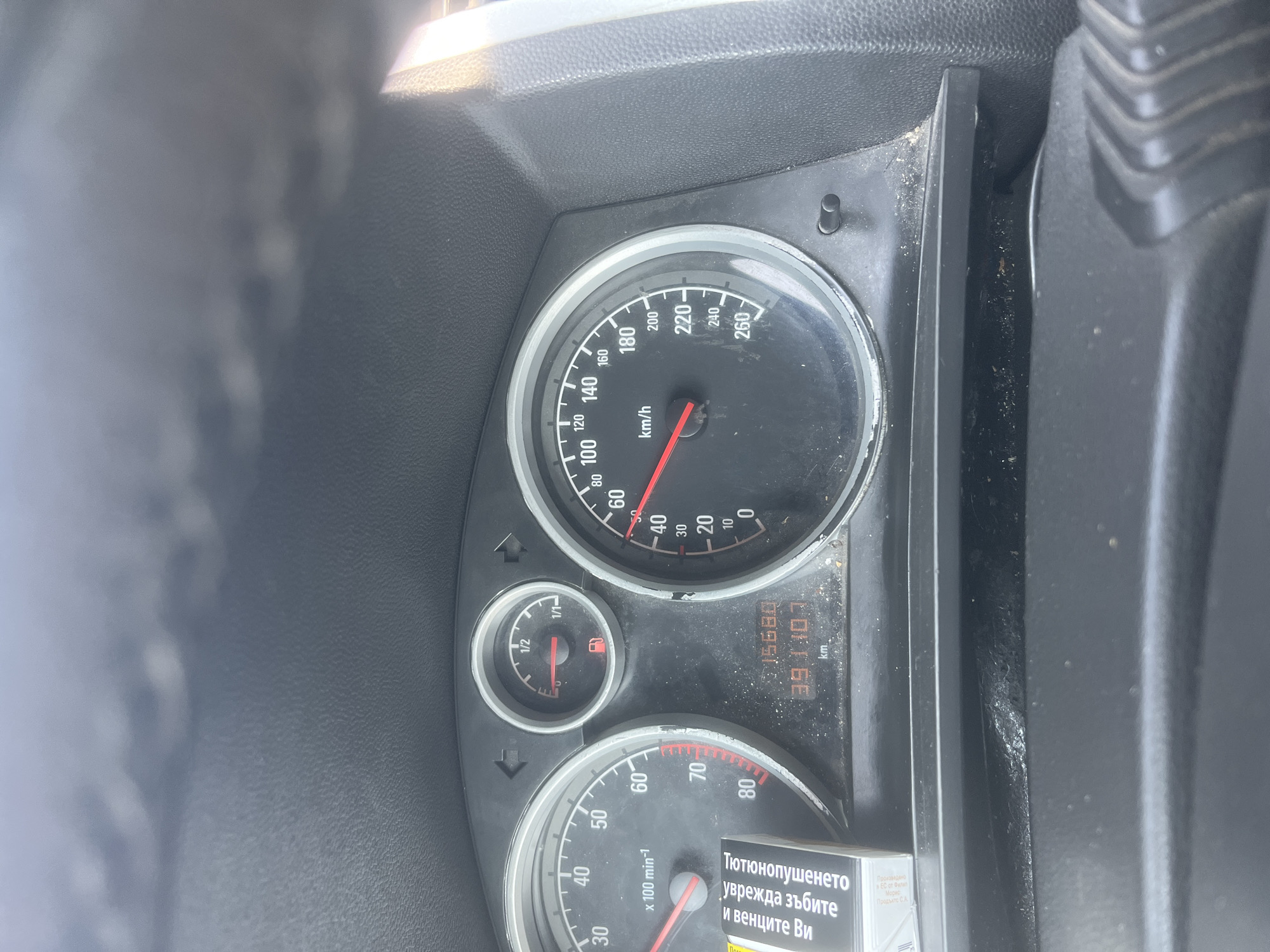The image displays the interior of a car, specifically focusing on the instrument cluster from the driver's perspective. The vehicle is stationary, indicated by the speedometer needle at 0 km/h (maximum 260 km/h) and the tachometer needle also at 0 x 100 min-1 (revolutions per minute, maximum 80 x 100 min-1 or 8000 RPM). The fuel gauge shows the tank is approximately one-quarter full.
An odometer display presents two numerical readings: "15680" and "391:10.7 km.". The first number likely represents the total distance traveled by the car, while the second, clearly marked with "km.", appears to be a trip odometer, possibly indicating 391.1 or 391.07 kilometers for a specific journey.
The dashboard and instrument panel show visible signs of dust and dirt accumulation. Bright sunlight is evident through reflections on the dashboard and windshield area, suggesting it is daytime.
In the lower right corner, a portion of a cigarette pack is visible, featuring a Bulgarian health warning: "Тютюнопушенето уврежда зъбите и венците Ви" (Smoking damages your teeth and gums). Additional text on the pack identifies it as "Произведено в ЕС от Филип Морис Продъктс С.А." (Produced in the EU by Philip Morris Products S.A.). The Cyrillic script and the brand information are consistent with the location context of Varna, Bulgaria. No people are visible in the frame.