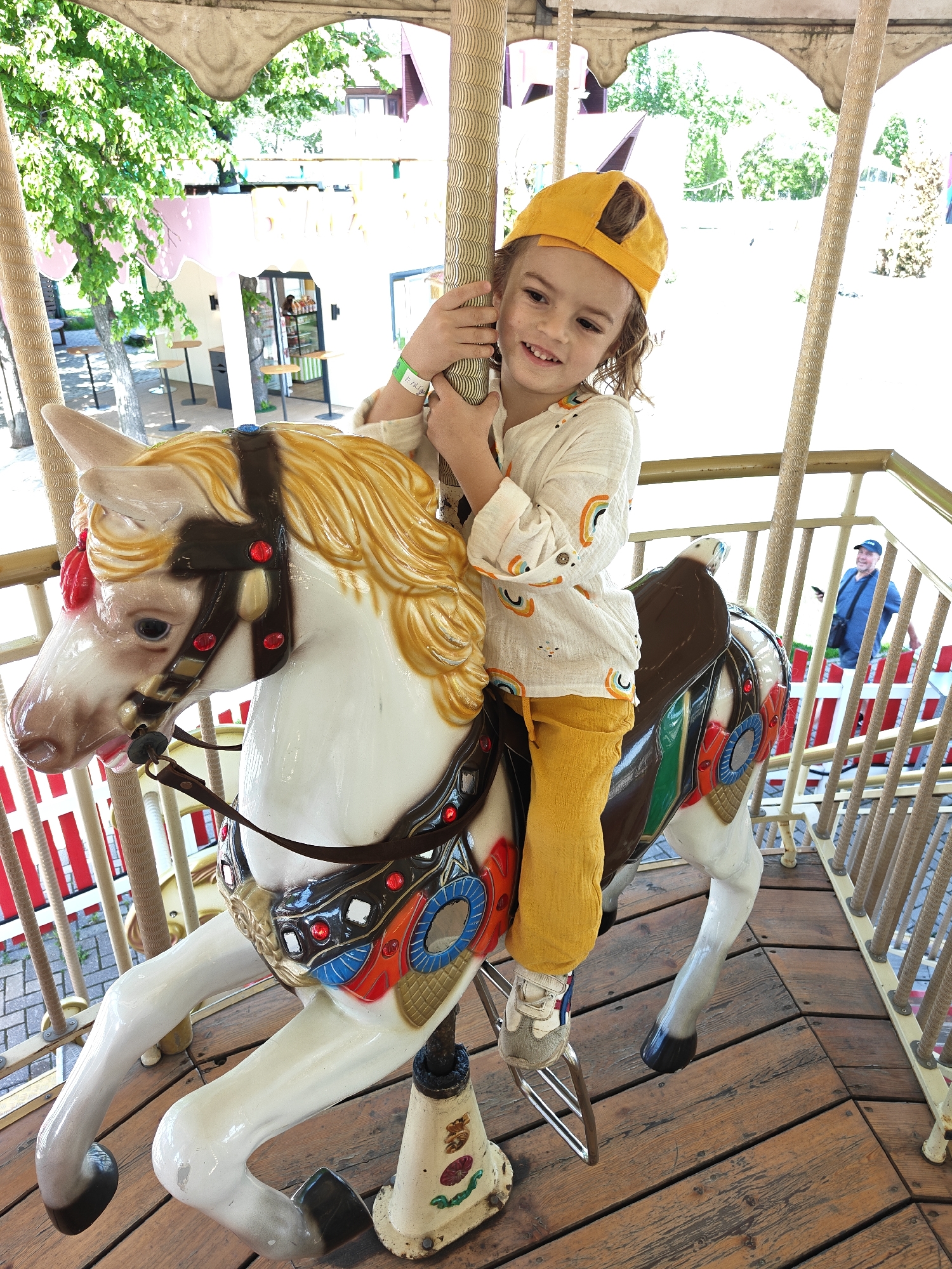 A young child in a yellow cap and clothing is riding a carousel horse in an outdoor setting. The carousel horse is white with a brown saddle and colorful decorations. The child is smiling and holding onto the pole. In the background, a man in a blue shirt and cap can be seen behind a railing. The location is Khimki, Russia.