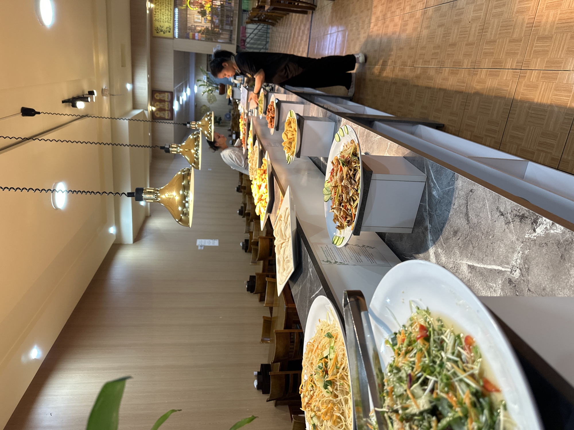 The image shows a buffet line in a restaurant. Several dishes are displayed on a long counter, including salads, noodles, and other food items. Two women, likely employees, are seen tending to the buffet. One woman, wearing black, is arranging food on a platter, while another woman, in a white top, is standing further down the counter. The setting appears to be indoors, with warm lighting from overhead fixtures illuminating the food and the dining area. The floor is tiled with a wood-like pattern, and the counter has a marble-like finish. In the background, rows of wooden chairs suggest a dining area. The overall atmosphere seems to be busy, indicative of a meal service in a restaurant. There is no visible text in the image.