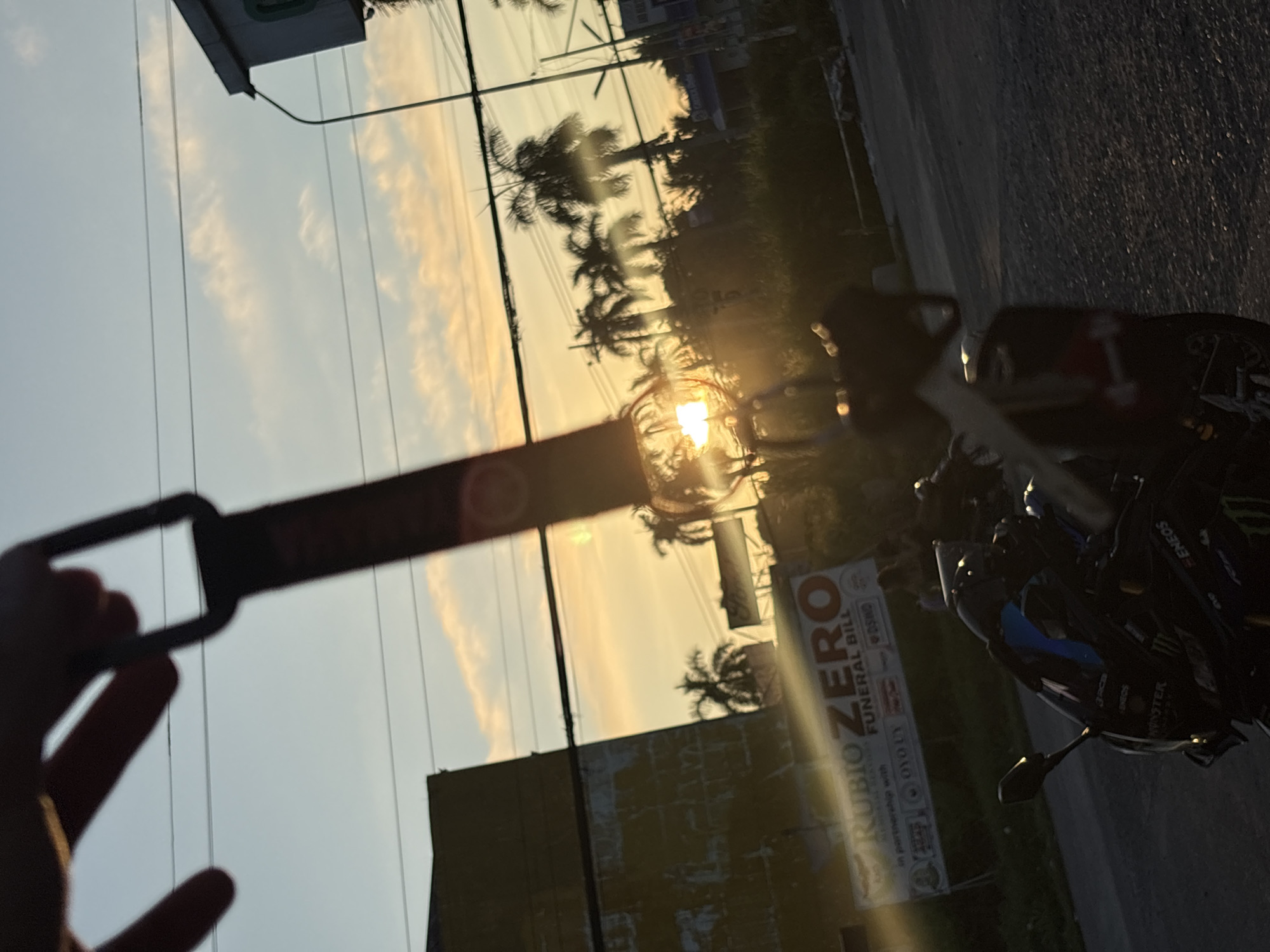 A person's hand holds up a dark keychain with a rectangular loop, obscuring a portion of a sunset over a street in Tagum City, Philippines. The sun, a bright orb, is partially visible through palm trees and is casting lens flares. In the foreground, a dark motorcycle with racing decals, including "MONSTER ENERGY" and "EN EOS", is parked. Behind the motorcycle, a banner with the text "RUBIO ZERO FUNERAL BILL" is visible. The background features power lines, a building with a yellow facade, and more palm trees. The sky is a mix of blue and orange, indicating either sunrise or sunset.