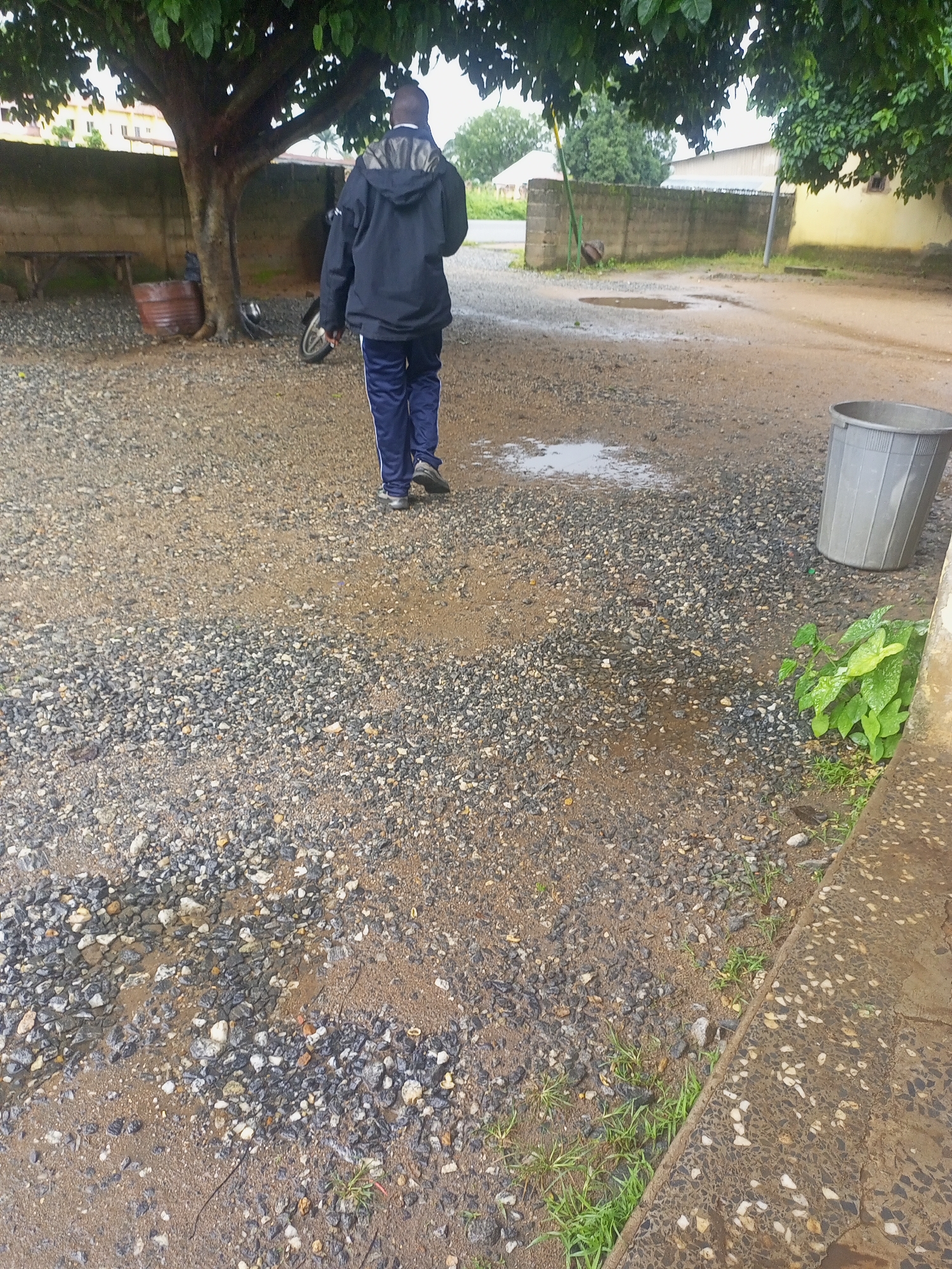 The image depicts a person walking away from the camera on a gravelly surface in what appears to be an outdoor courtyard. The person is wearing a dark blue jacket and matching trousers with white stripes down the sides. The background shows a wall, a tree, and some buildings. A large gray trash bin is visible to the right of the frame. There are puddles of water on the ground, suggesting it may have recently rained. The overall environment appears to be a somewhat utilitarian or residential area. The scene is set in Serti, Nigeria.