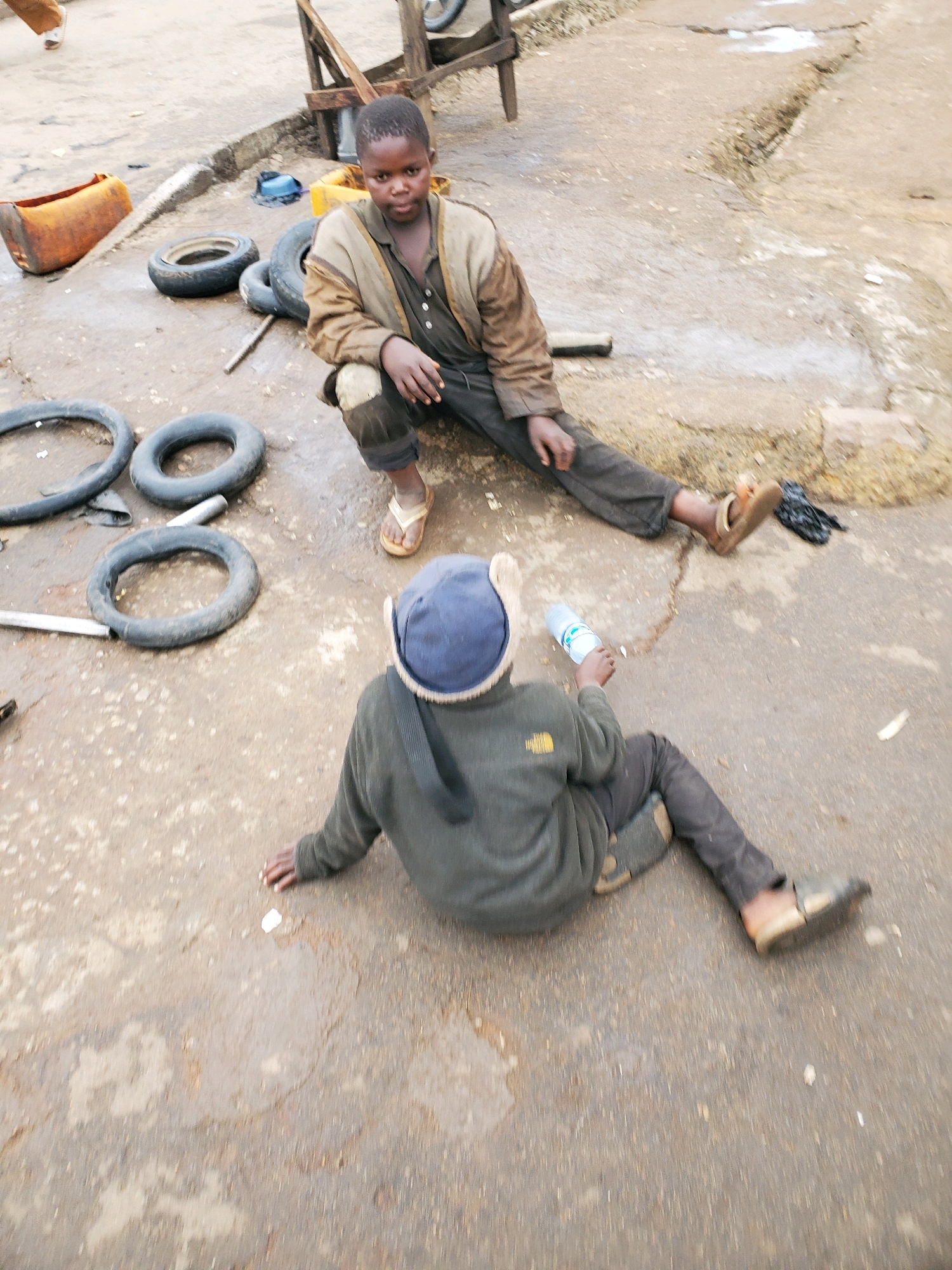 Two young boys are sitting on a wet, cracked asphalt ground in what appears to be an outdoor workshop or scrap yard. The boy in the foreground, wearing a dark green hooded jacket and dark pants, is facing away from the camera, holding a clear plastic water bottle in his right hand. He has a blue beanie on his head and is wearing dark shoes. The boy in the background, facing the camera, is sitting with his legs extended. He is wearing a light brown jacket over a dark shirt and dark pants, with his knees exposed. He has on light-colored sandals. Several used tires are scattered around them on the ground, some are partially submerged in puddles of water. To the left of the image, an orange, cylindrical object, possibly a fuel container, rests on the ground. In the background, a wooden structure, possibly part of a workbench or shelter, is visible. The overall environment suggests a utilitarian area, likely within the city of Jos, Nigeria, given the context. The scene depicts a moment of rest or pause for the two boys amidst their surroundings.