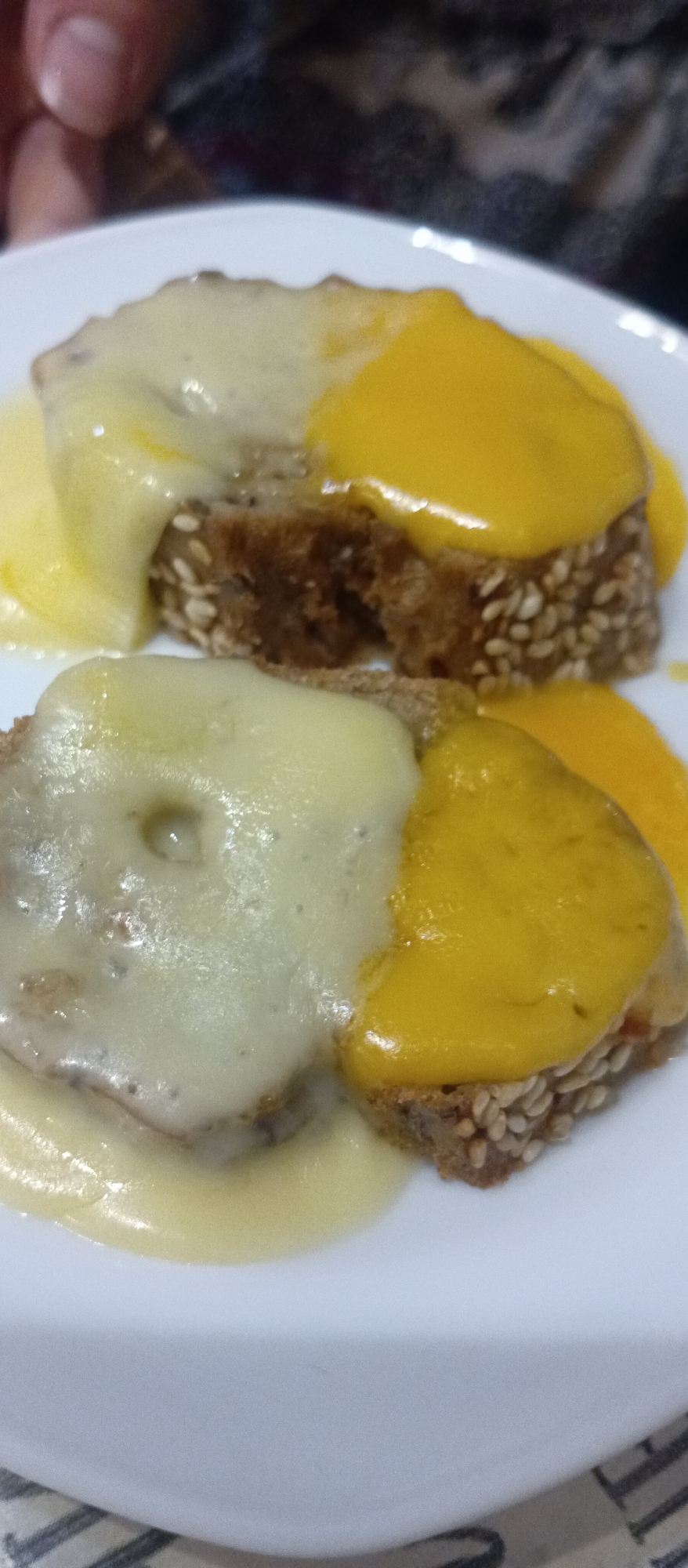 On a white plate, two pieces of sesame-seed bread are each topped with two different types of melted cheese: one side with a pale yellow, creamy cheese and the other side with a bright yellow cheese. Melted cheese has flowed over the edges of the bread. A finger is visible in the upper-left corner of the image. The specific location and city are unknown.
