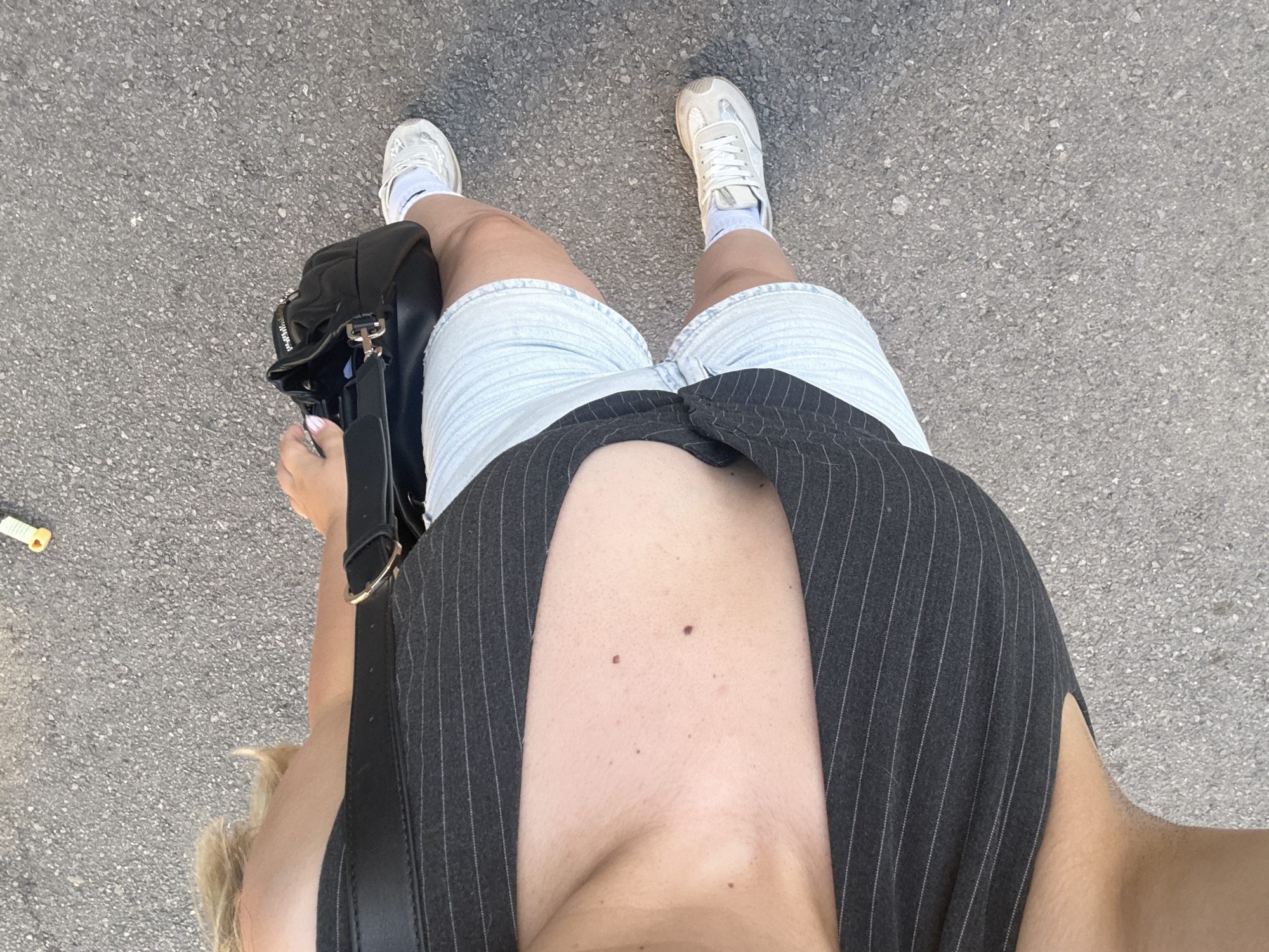 A woman stands on asphalt in Sofia, Bulgaria. She wears light blue denim shorts, a black pinstriped vest, white socks, and white sneakers. A black leather purse hangs from her shoulder. A discarded yellow wrapper lies on the ground to the left.