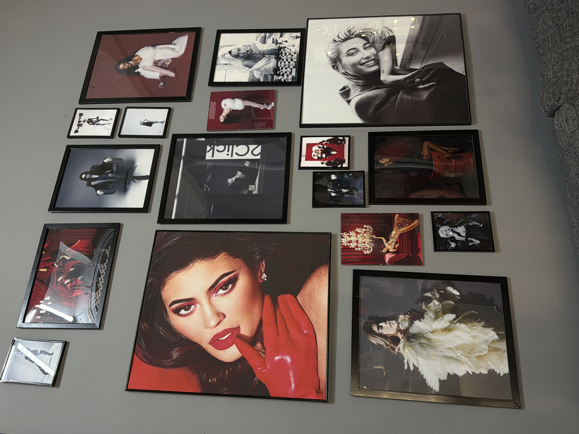 A wall is adorned with 16 framed pictures of various shapes and sizes. The frames are predominantly black, with one silver frame and several small unframed prints. The subjects of the pictures range from portraits of women to abstract designs. The wall is gray. The location and city of Kazan, Russia are not evident from the image.