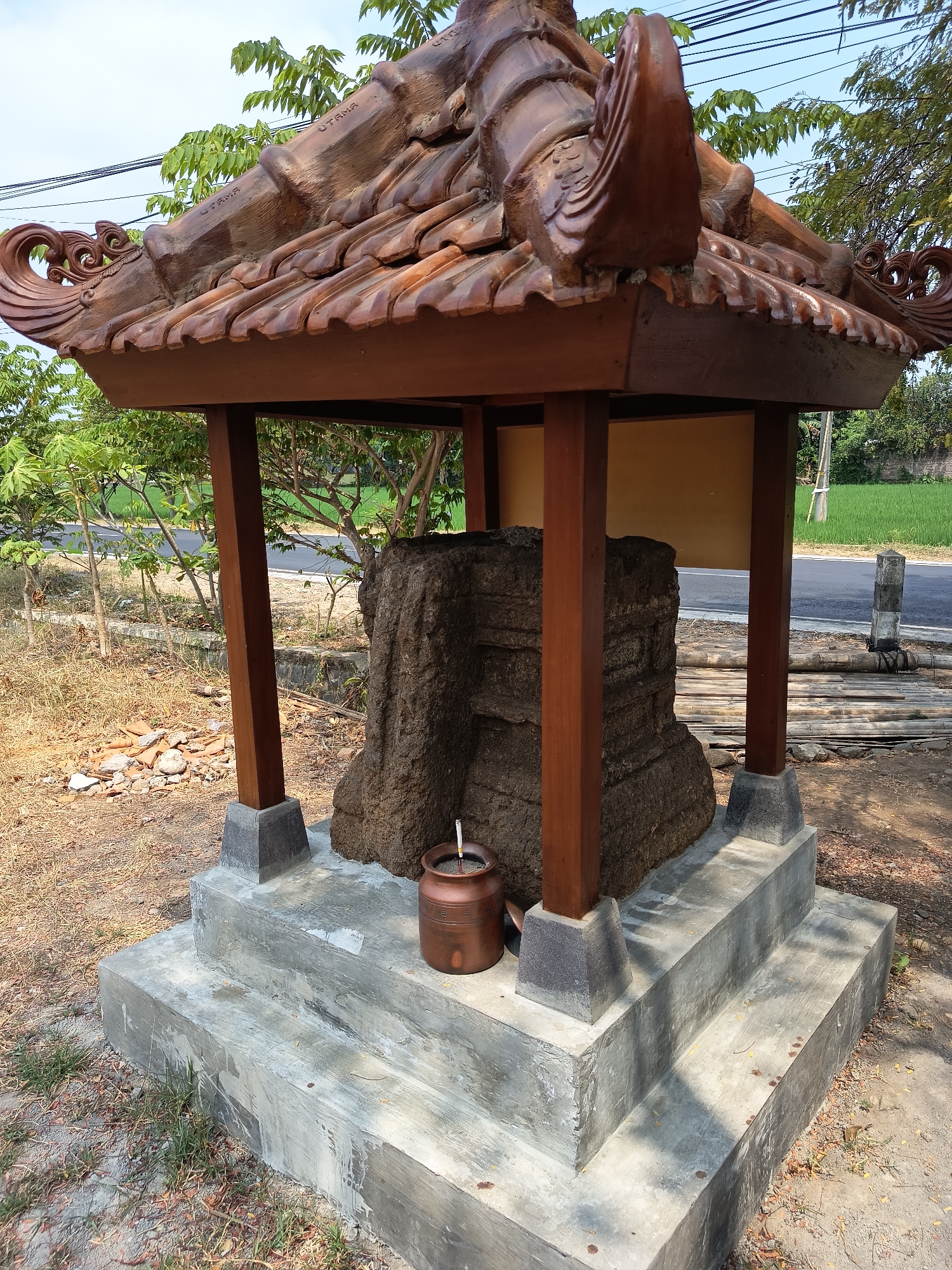 In Lalung, Indonesia, a stone monument, possibly a relic from an ancient civilization, stands under a wooden gazebo with a tiled roof. The stone is rough-hewn and appears to be layered, with some sections showing carved patterns. A small, ornate clay pot, possibly for incense, sits on the stone steps leading up to the monument. The gazebo is supported by four wooden posts, each resting on a concrete base. The surrounding environment is dry and dusty, with sparse vegetation and a paved road visible in the background. The sky is clear and blue, indicating a sunny day. The scene suggests a historical site or a place of cultural significance.