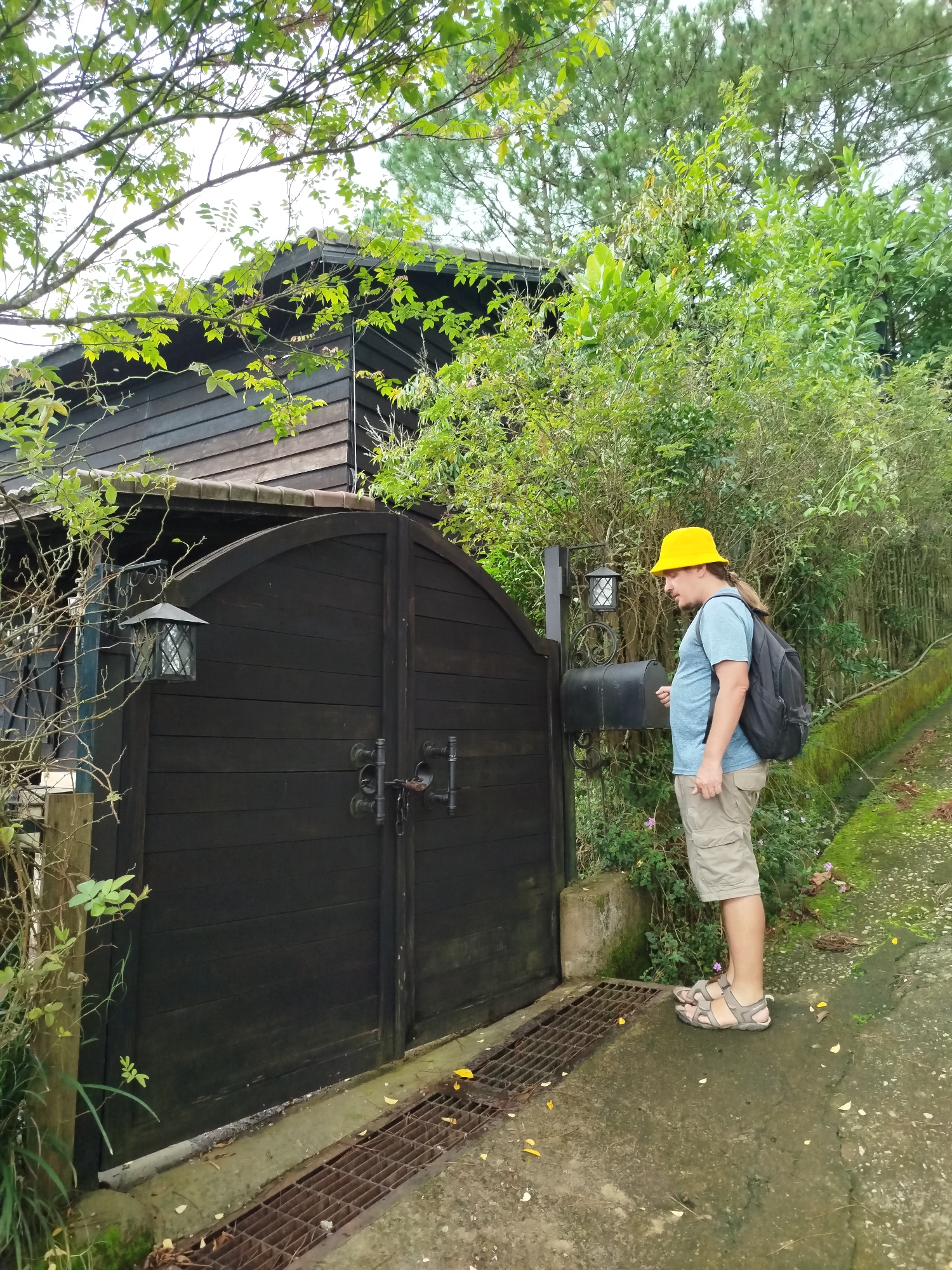A man wearing a yellow bucket hat, a light blue t-shirt, and khaki shorts stands on a moss-covered path, looking at a black mailbox next to a dark wooden gate. He has a black backpack on. The gate is adorned with decorative metal handles and a latch. A lantern hangs on a post to the left of the gate, and another lantern is visible on the mailbox post. The background features a rustic wooden house with a dark roof, overgrown with lush green trees and foliage, suggesting a natural, possibly rural or semi-rural setting within Cam Ly - Da Lat, Vietnam. A rusty metal grate is embedded in the concrete path in front of the gate, and fallen yellow leaves are scattered around.
