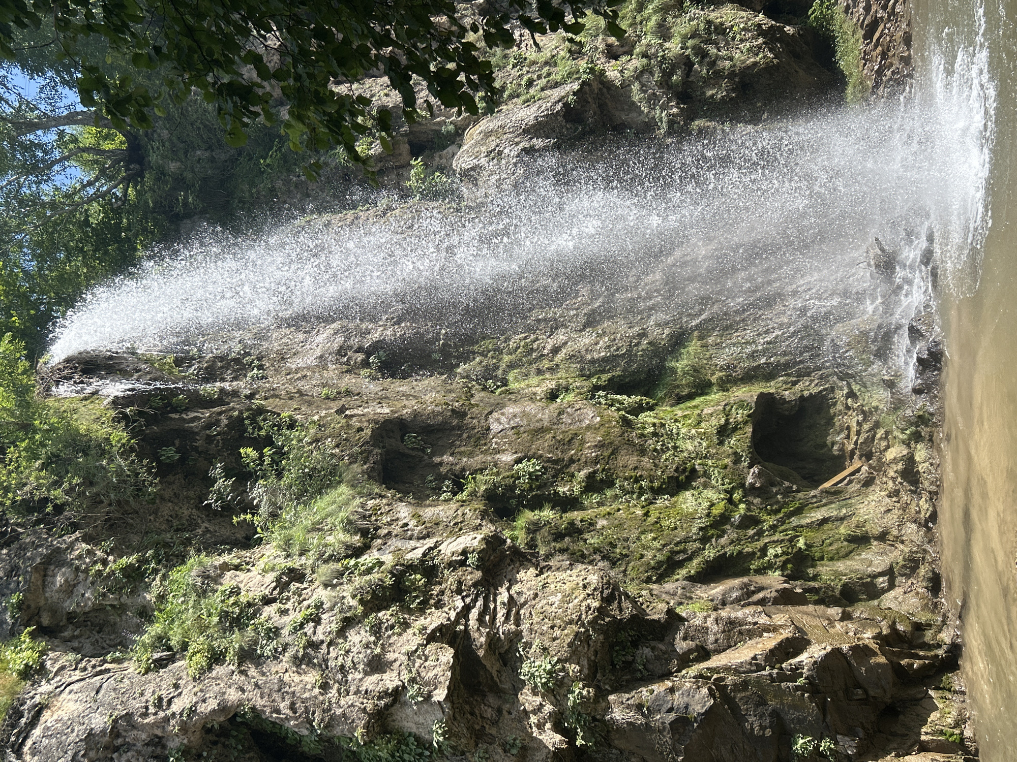 The image shows a waterfall cascading down a rocky cliff face covered in moss and vegetation. The water falls into a shallow pool at the base of the cliff. Trees and foliage are visible at the top of the cliff and on its sides. The lighting suggests it is a sunny day. The location is identified as Krasny Kurgan, Russia, though this specific waterfall is not a widely recognized landmark. There are no people or animals visible in the image.