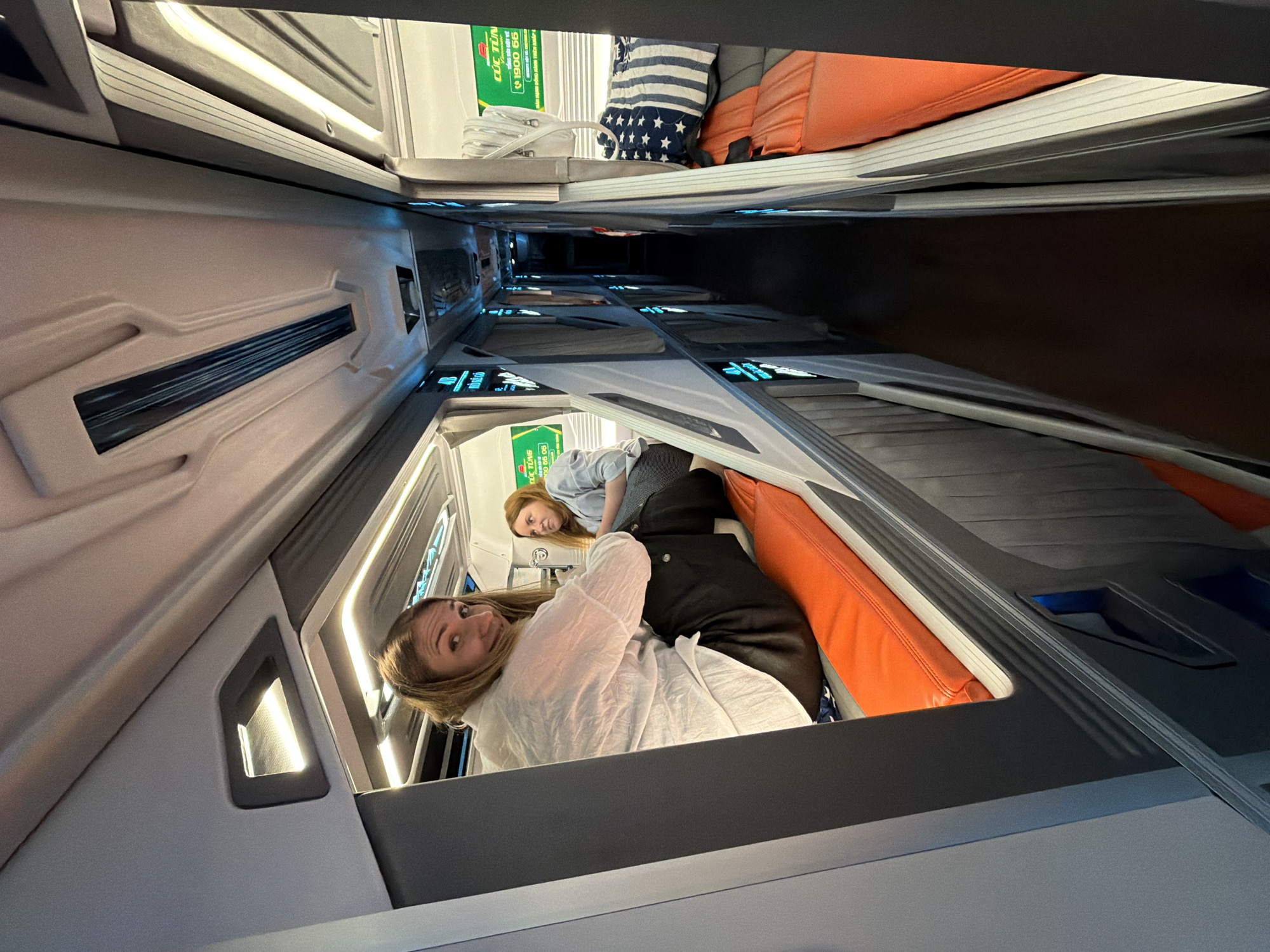 In Đại Lãnh, Vietnam, two young women are seated in a sleeper bus. The woman in the foreground, with blonde hair, wears a white shirt and black pants. She is looking at the camera and sitting on an orange cushion. To her right, another woman with red hair, wearing a blue shirt, is also seated on an orange cushion. The bus has narrow sleeping compartments, with blue lighting and luggage visible in the background. The interior design is modern and functional, with a focus on maximizing space for passengers. The overall atmosphere appears to be relaxed and casual, suggesting a journey or stopover.