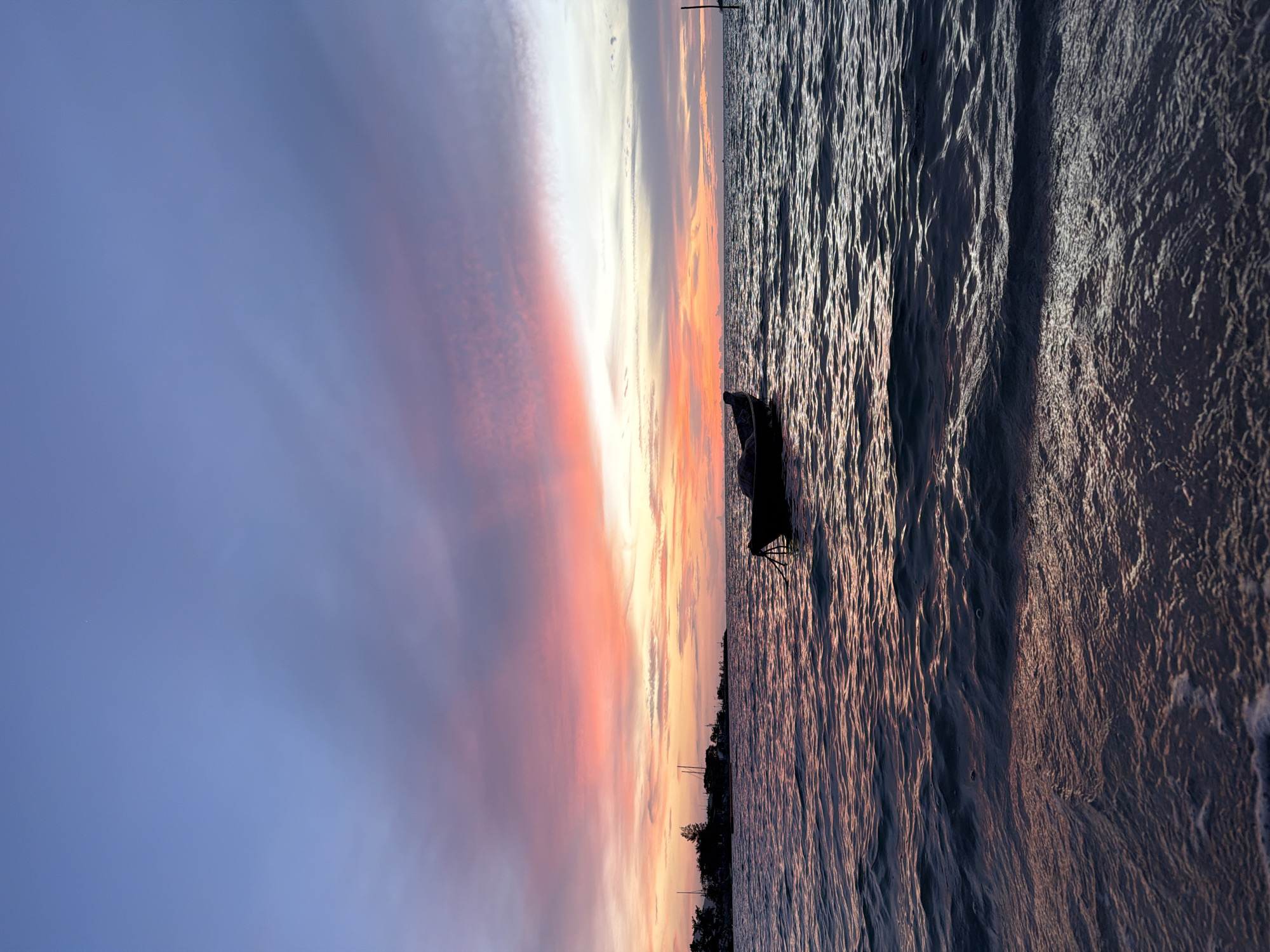 A serene sunset is depicted over the ocean in Ko Samui District, Thailand. The sky is a gradient of colors, transitioning from a deep blue at the top to vibrant pinks and oranges near the horizon. Wispy clouds catch the light, creating streaks of color across the sky. In the middle ground, a small, dark silhouette of a boat floats on the calm water, tethered by a rope. The water itself reflects the colors of the sunset, with gentle waves lapping at the shore in the foreground. Along the distant horizon, a dark silhouette of a landmass with trees and structures can be seen. The overall impression is one of peace and tranquility.