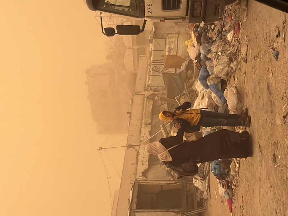 The image depicts an outdoor urban environment engulfed in a dense, orange-brown dust storm, significantly reducing visibility. The specific location within the city of Unknown, Unknown is indeterminate, but the scene suggests an impoverished or damaged area, possibly a refugee camp or neglected urban periphery.
In the foreground, the ground consists of unpaved dirt and scattered debris. Two individuals are positioned slightly left of center. The person on the left, an adult, wears dark long clothing and a light-colored headscarf, with their back partially turned towards the viewer. They carry a dark backpack on their left shoulder and their left arm is extended towards the second individual. The second individual, a child, wears a yellow hooded jacket, blue jeans, and brown boots, also carrying a backpack. The child faces the adult, with their left hand raised towards their face/neck.
To the immediate left of the individuals stands a light-colored, rudimentary structure with an open doorway. Dominating the midground between the individuals and the background is an extensive accumulation of garbage, debris, and bundled items, some wrapped in blue material. To the far right, the front portion of a large, light-colored truck is partially visible, displaying the number "276" on its side. In the background, obscured by the heavy dust, are outlines of multiple dilapidated or incomplete buildings and overhead utility lines connected to poles. The entire scene is bathed in a uniform orange hue from the dust-laden atmosphere.