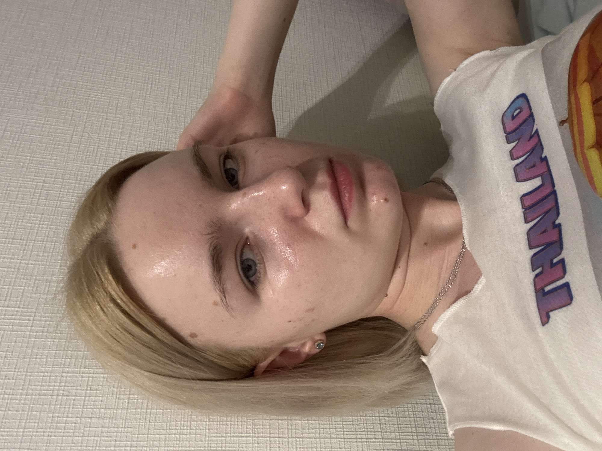 A young woman with blonde hair and blue eyes is pictured, looking directly at the camera. She has several moles on her face and a small dark mole on her chin. She is wearing a white tank top with the word "THAILAND" printed on it in colorful lettering and a silver chain necklace. Her left arm is raised and resting on her head. The background appears to be a textured, light-colored wall. The image is a close-up shot, focusing on the woman's upper body and face. There is no indication of location in the city of Unknown, Russia.