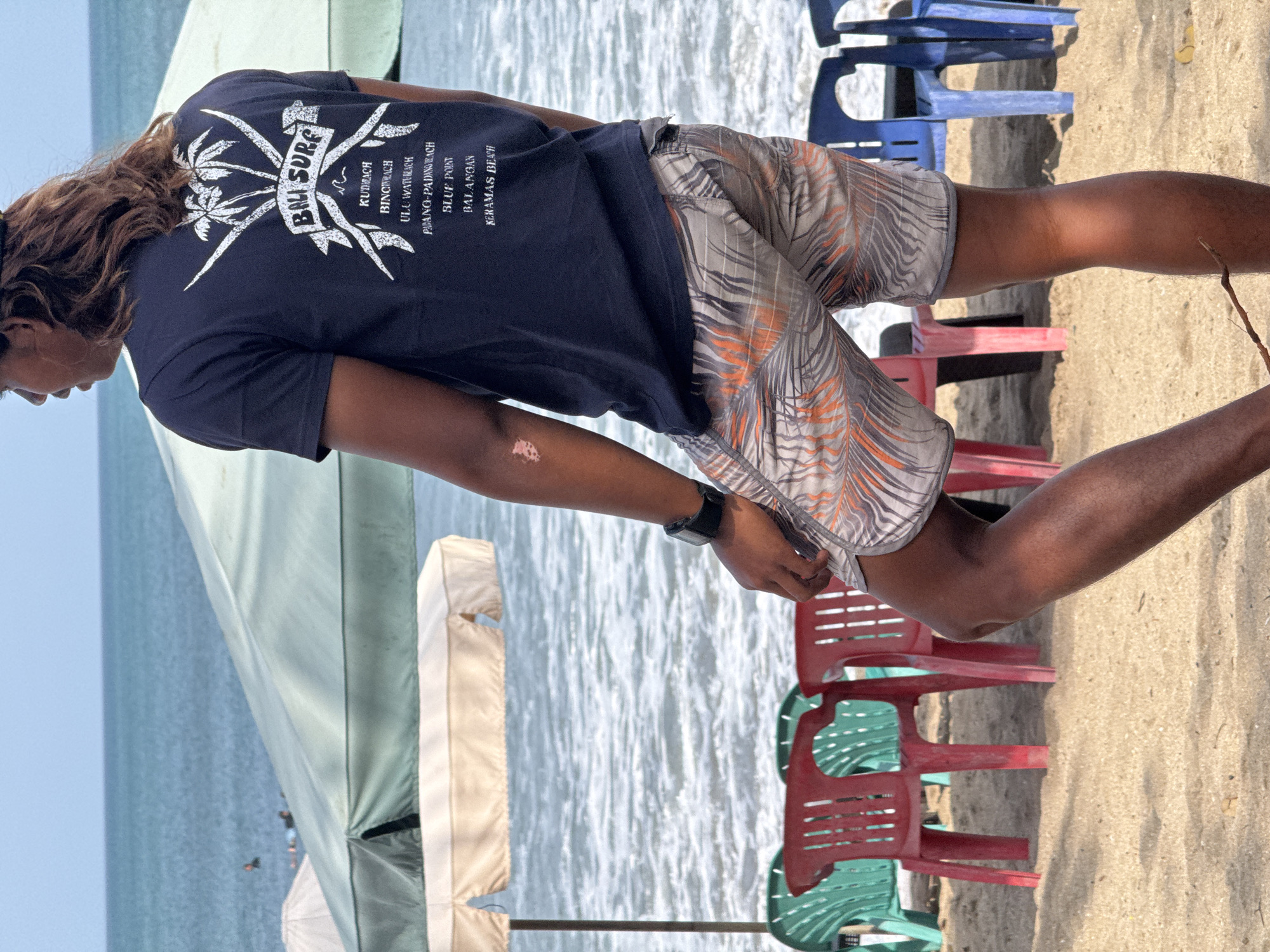 A person is standing on a sandy beach in Kuta, Indonesia. They are wearing a dark blue t-shirt with a white graphic design on the back that says "BALI SURF" and lists several beach names. They are also wearing patterned shorts. The person is looking out at the ocean, which is visible in the background, with waves. There are several beach chairs, some green, some red, and some blue, set up on the sand behind the person. Two large umbrellas, one white and one green, are also present on the beach. The sky is clear and blue. The person has long, dark hair. A portion of their left arm shows vitiligo.