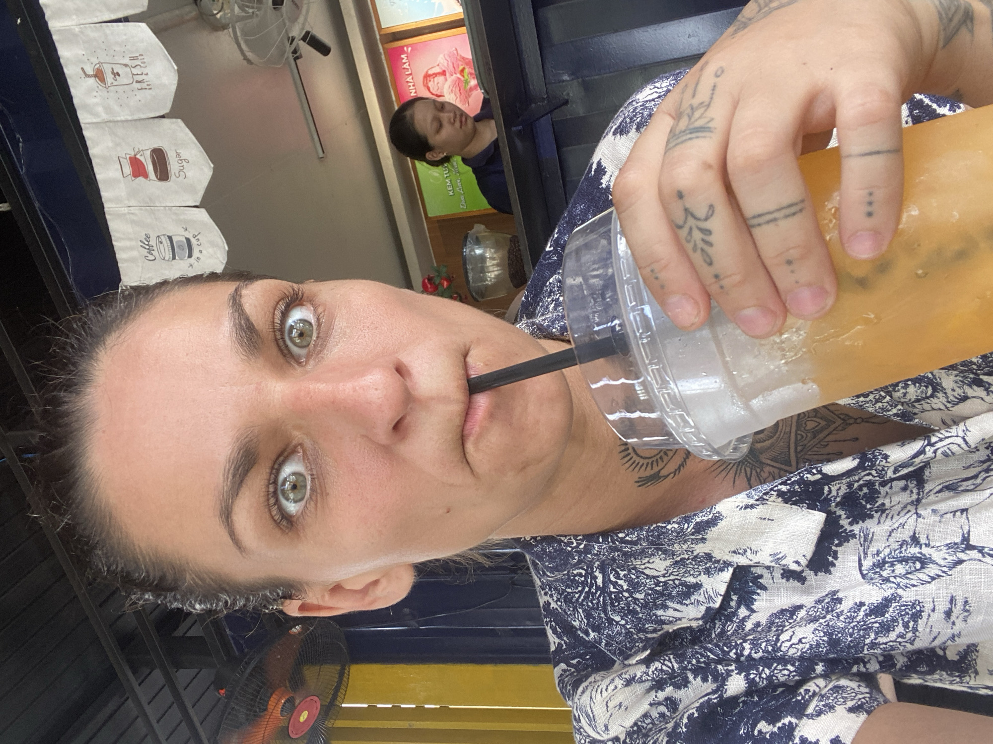 The image is an eye-level, rotated selfie of a person with light skin, green eyes, and dark hair, drinking from a clear plastic cup with a black straw. Their face is close to the camera, and their gaze is directed at the viewer. They have visible tattoos on their hand and neck. The person is wearing a blue and white patterned shirt. In the background, another person with dark hair is visible. Coffee themed decor hangs in the background. An electric fan is also visible. The location is in the city of Đồng Nai, Vietnam.