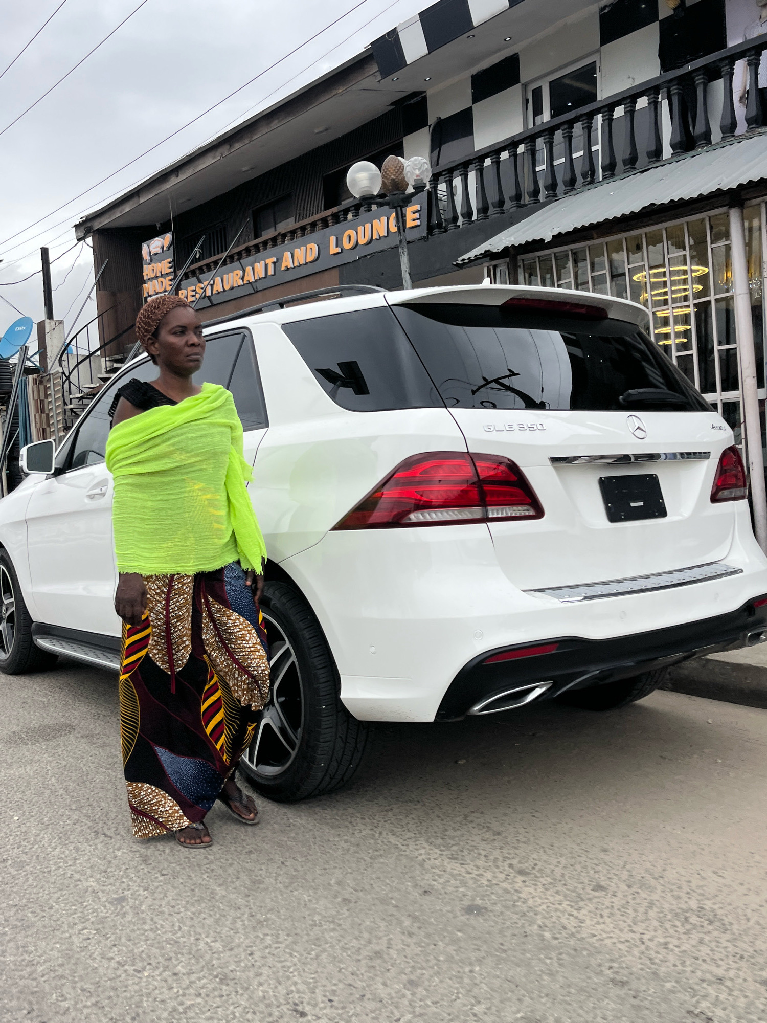 A woman stands next to a white Mercedes-Benz GLE 350 in front of a two-story building that houses a restaurant and lounge. The building has a black and white checkered facade on the upper level and a dark brown facade on the lower level. A sign above the lower level reads "HOME MADE RESTAURANT AND LOUNGE" in orange lettering. The woman is wearing a bright yellow wrap over a long, colorful patterned skirt and dark sandals. She is looking to her left. The car is parked on a paved street. The sky is overcast. The scene appears to be in Ado, Nigeria.