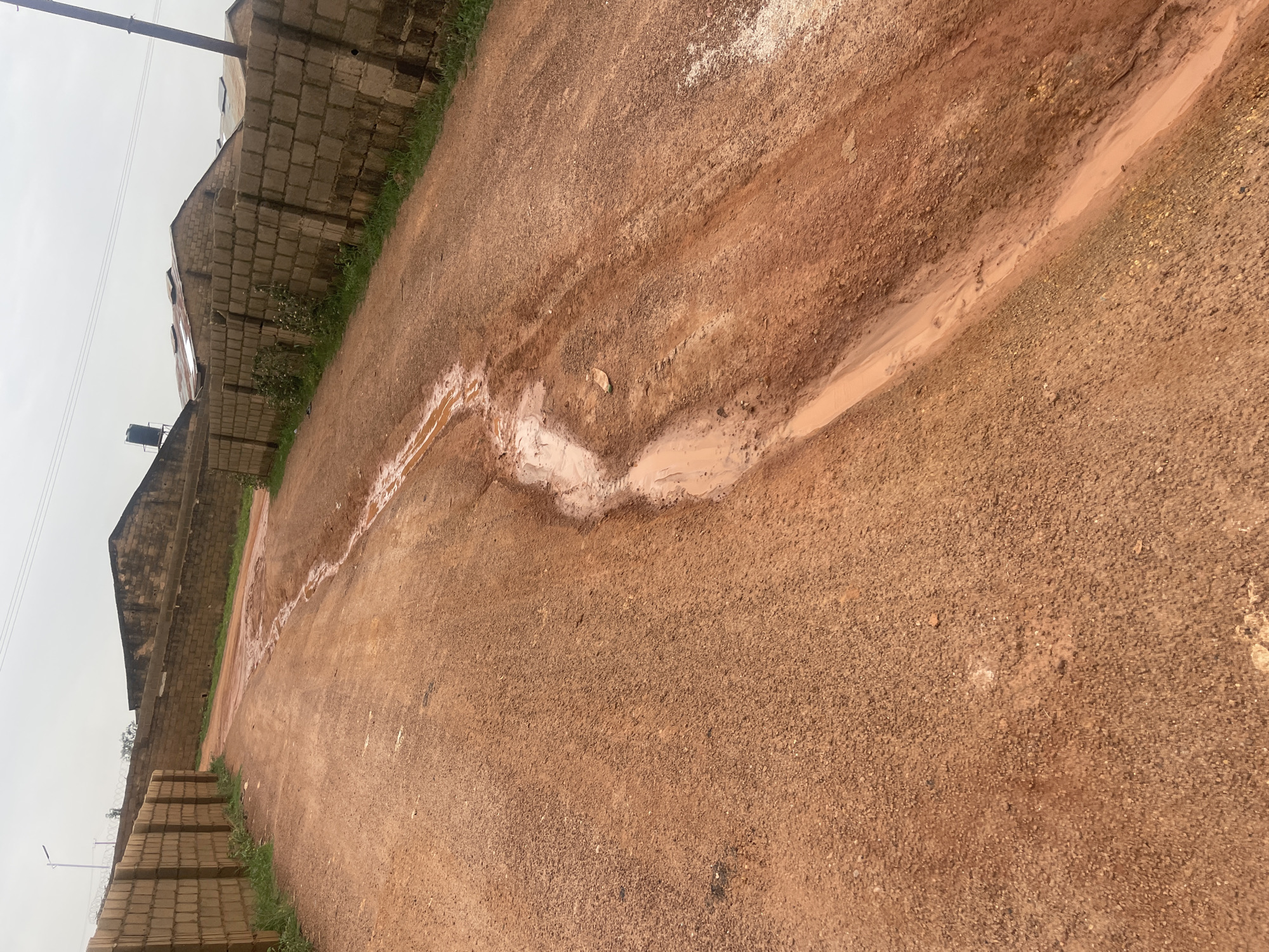 This media file depicts an outdoor scene in Jos, Nigeria. The primary subject is a dirt road with a muddy, water-filled ditch running alongside it. The road is made of reddish-brown dirt and gravel. To the left of the road, there is a low wall constructed from concrete blocks. Beyond this wall, structures are visible, including buildings with corrugated metal roofs and a water tank. Further to the left, stacks of concrete blocks are piled on the side of the road, adjacent to a thin strip of green grass. A power line is also visible running horizontally across the sky. The sky is overcast and light, suggesting a cloudy or possibly rainy day. The overall scene conveys a sense of a developing or undeveloped area, common in many parts of Nigeria. There is no visible text in the image. The lighting suggests it is daytime, but the overcast sky makes it difficult to determine the exact time. The presence of the muddy ditch indicates recent rainfall or poor drainage.