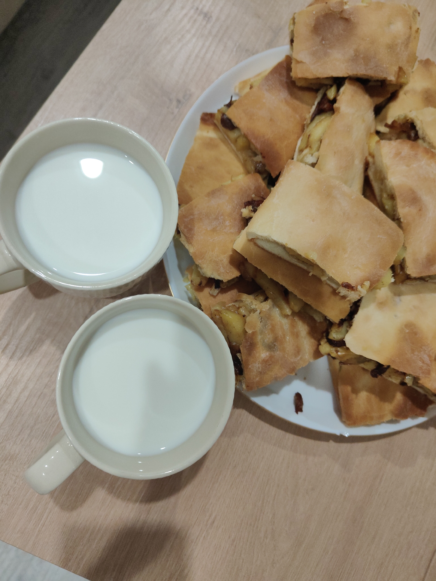 On a light brown wooden surface are two beige mugs filled with white liquid, likely milk, and a white plate piled with square-cut pieces of baked pastry. The pastry appears to have a filling. The scene is indoors. The location is Unknown, Unknown.