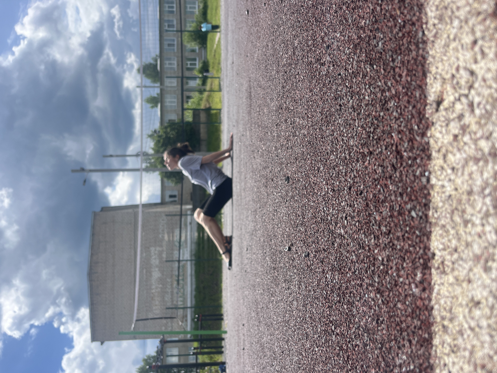 An adult, with dark hair pulled back, is lying on their back with their legs extended on a reddish-brown asphalt surface. They are wearing a grey t-shirt and black leggings with dark sandals. Their head is tilted back and looking upwards towards a cloudy sky. Behind the person, a white volleyball net is stretched across a court. Further in the background, a light-colored brick building with multiple windows stands to the left, and a row of multi-story buildings with a light facade and many windows is visible to the right. A street light pole is present on the left side of the frame. In the far right background, a person in a light blue shirt is standing near a fence. The environment appears to be an outdoor sports or recreational area.