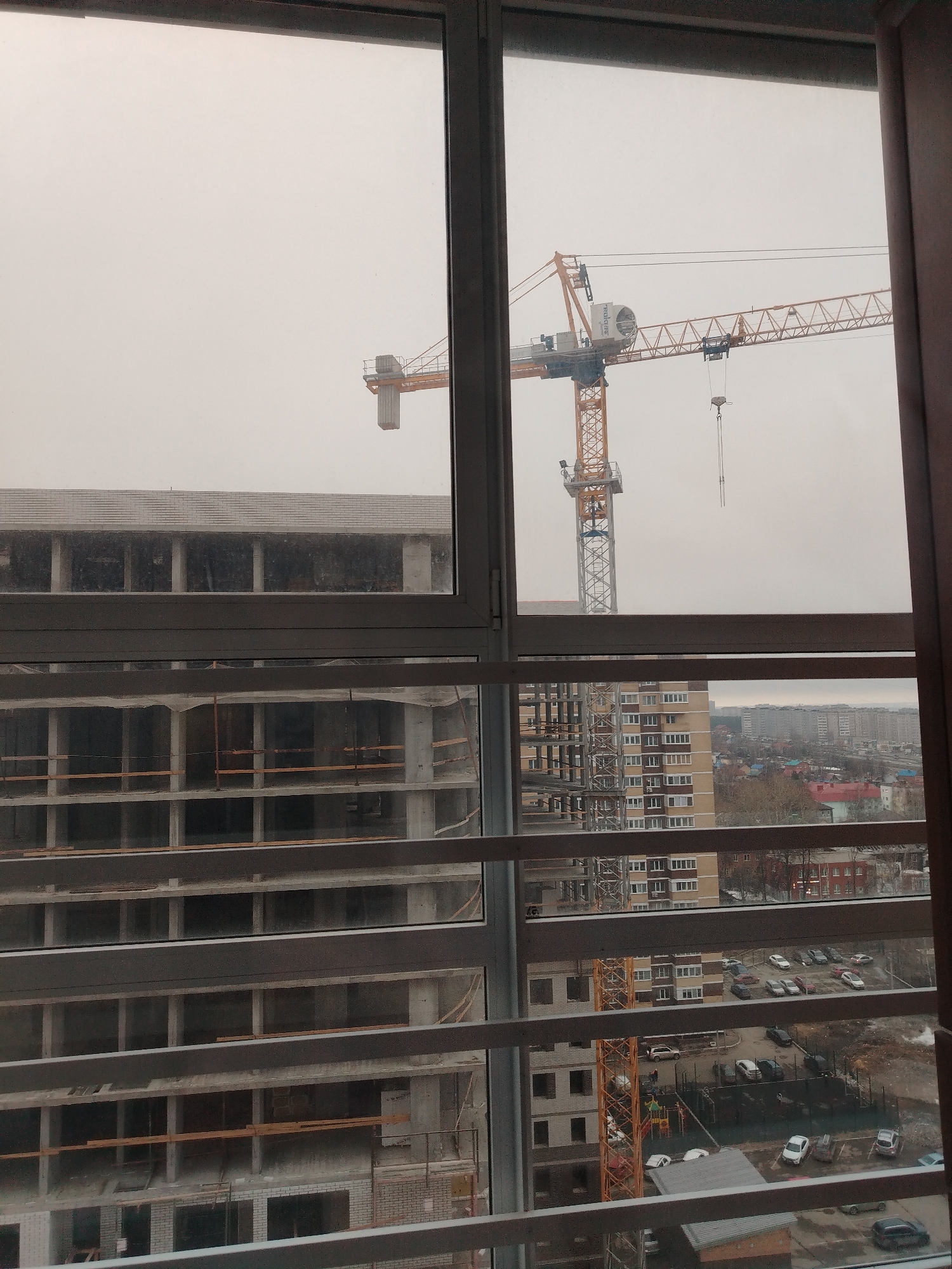 The image, likely taken from inside a high-rise building in Izhevsk, Russia, presents an overcast urban scene. A large construction crane dominates the view, positioned near a building under construction with an unfinished concrete structure. The city skyline is visible in the background, with multiple residential buildings and trees. A parking lot with various vehicles is also visible near the construction site. The image is framed by the windows of the building where the photo was taken, with their horizontal slats partially obstructing the view.