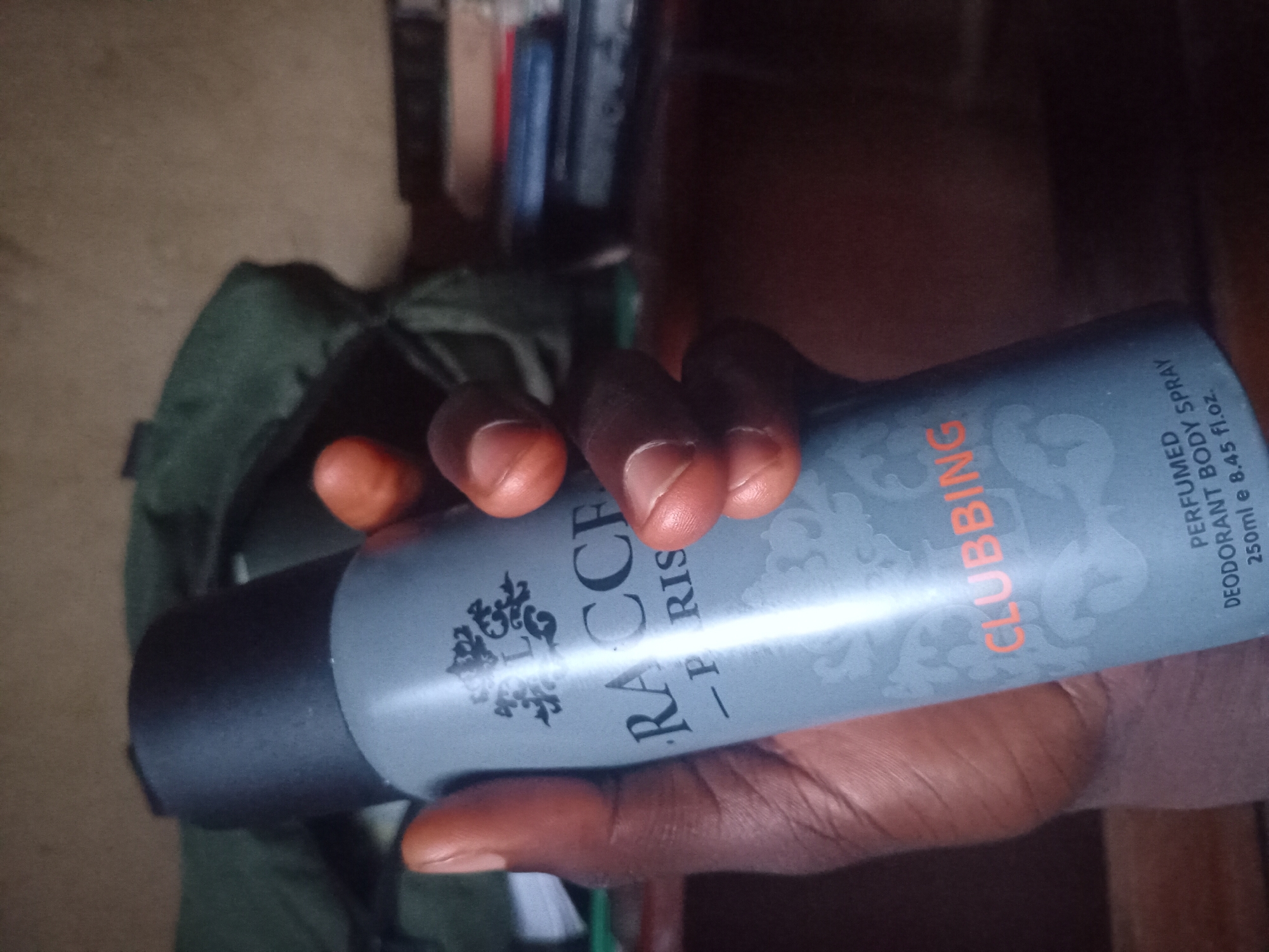 A person's hand holds a gray aerosol can of "RACCE PARIS CLUBBING PERFUMED BODY SPRAY 250ml e 8.45 fl.oz." The background is out of focus and appears to be an indoor setting with a green bag and some books on a shelf. The lighting is dim. The specific location in Bukuru, Nigeria, is not determinable from the image.