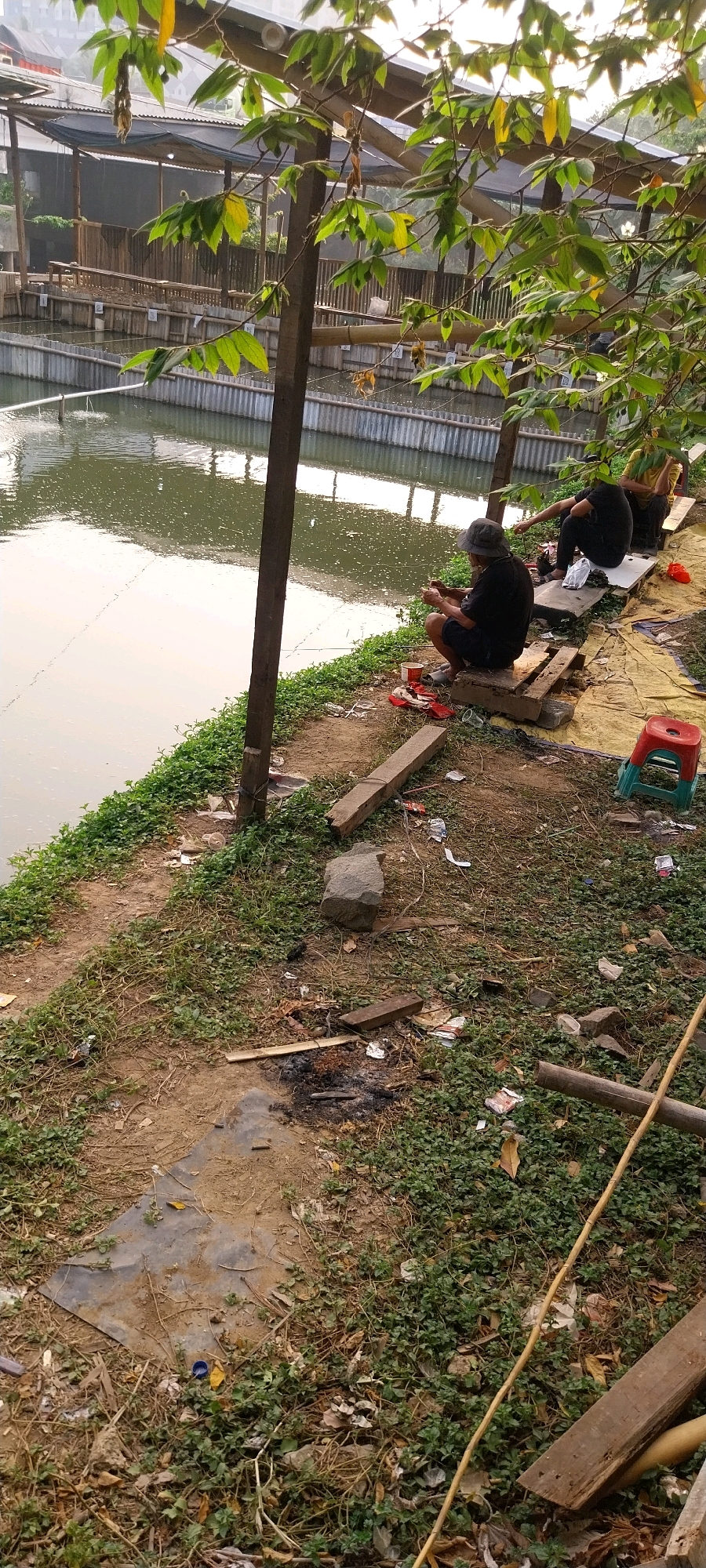 The image depicts an outdoor scene along a body of water, likely a canal or pond, in Tangerang, Indonesia. Three individuals are visible, all appearing to be male, engaged in fishing. They are seated on makeshift benches or platforms along the water's edge.
The individual in the foreground is wearing a dark shirt and shorts and a hat, holding a fishing rod. He is seated on wooden planks. To his right, another individual in a dark shirt and shorts is also holding a fishing rod. Further to the right, a third individual in a yellow shirt is seated, also with a fishing rod.
The environment is somewhat unkempt, with vegetation growing around the water's edge. Various objects are scattered on the ground, including plastic bottles, wrappers, and discarded materials, suggesting a casual and possibly frequented spot. A small red and green plastic stool is also visible. The water body has a calm surface with some ripples, and there appears to be some sort of fencing or enclosure bordering it in the background. Trees with green leaves overhang the scene, providing some shade. The overall impression is of a simple, informal fishing spot.