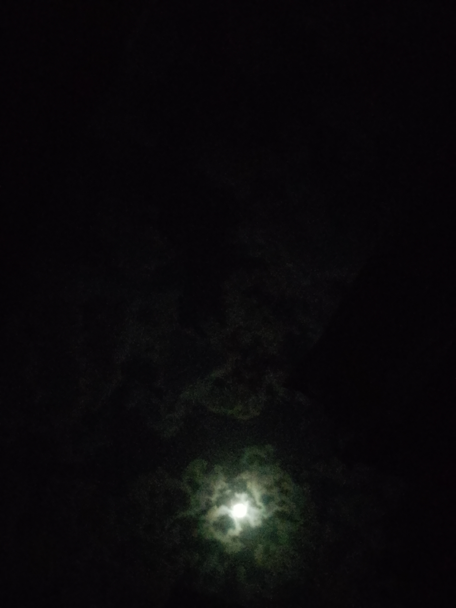 A low-angle, nighttime view reveals a bright, obscured light source, likely the moon, amidst a field of dark clouds. The light source, positioned near the bottom center of the frame, emits a diffused glow, blurring its distinct shape. The dark clouds surrounding the light source obscure the sky, and form shapes in the darkness. No people or objects are visible other than the moon and clouds. Location: Unknown, Unknown.