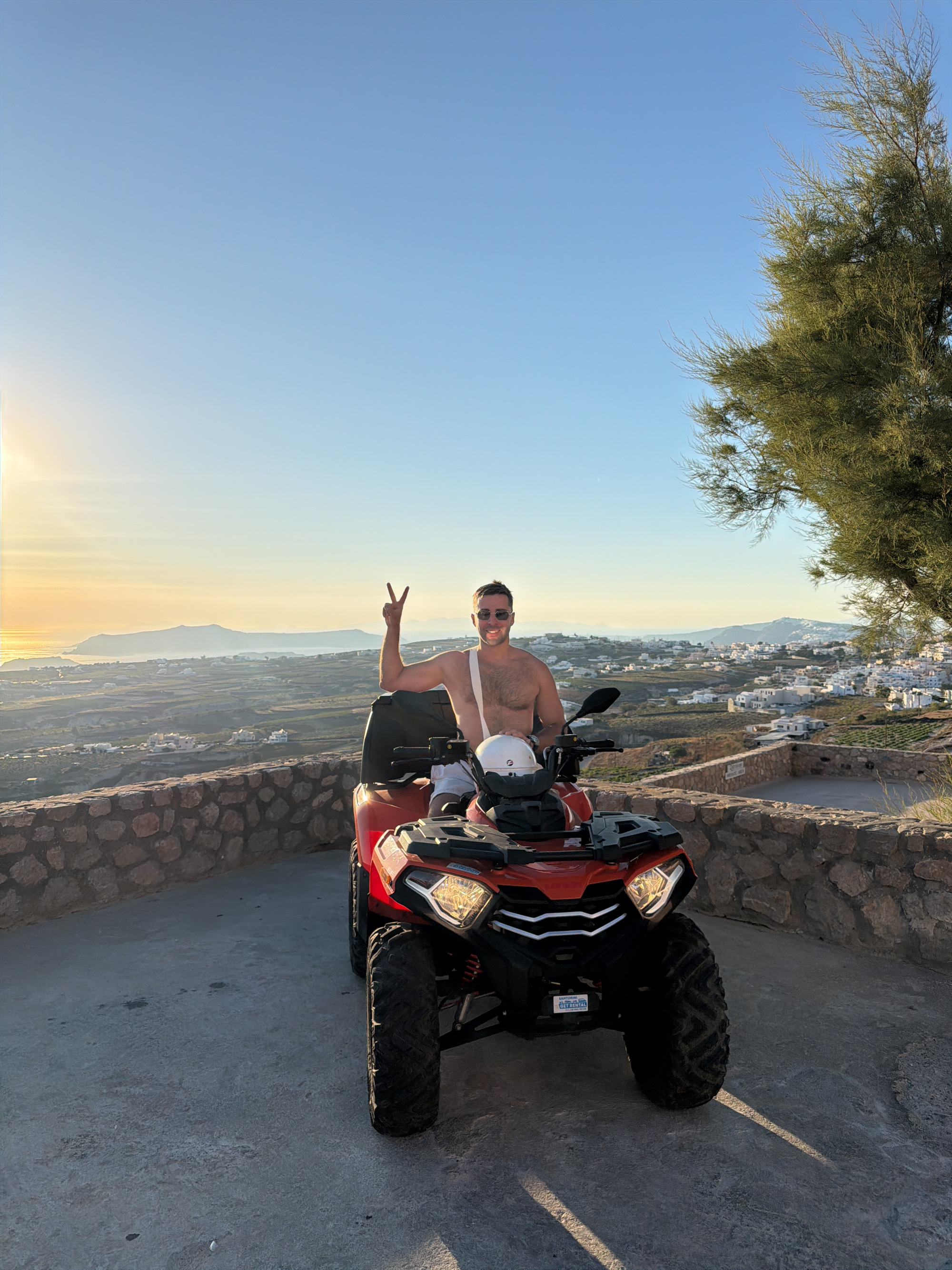 A young adult Caucasian male with short brown hair, wearing sunglasses, is sitting on a red ATV, giving a peace sign with his left hand raised. He is shirtless and appears to be wearing white shorts. A white helmet rests on the handlebars of the ATV. The ATV is parked on a concrete surface in front of a stone wall. In the background, a town with white buildings is visible on a hillside, overlooking the sea. The sun is setting, casting a warm glow over the landscape. A large tree with green foliage is positioned to the right of the ATV. The image appears to have been taken in a scenic location, possibly Santorini, given the architectural style of the town and the coastal landscape.