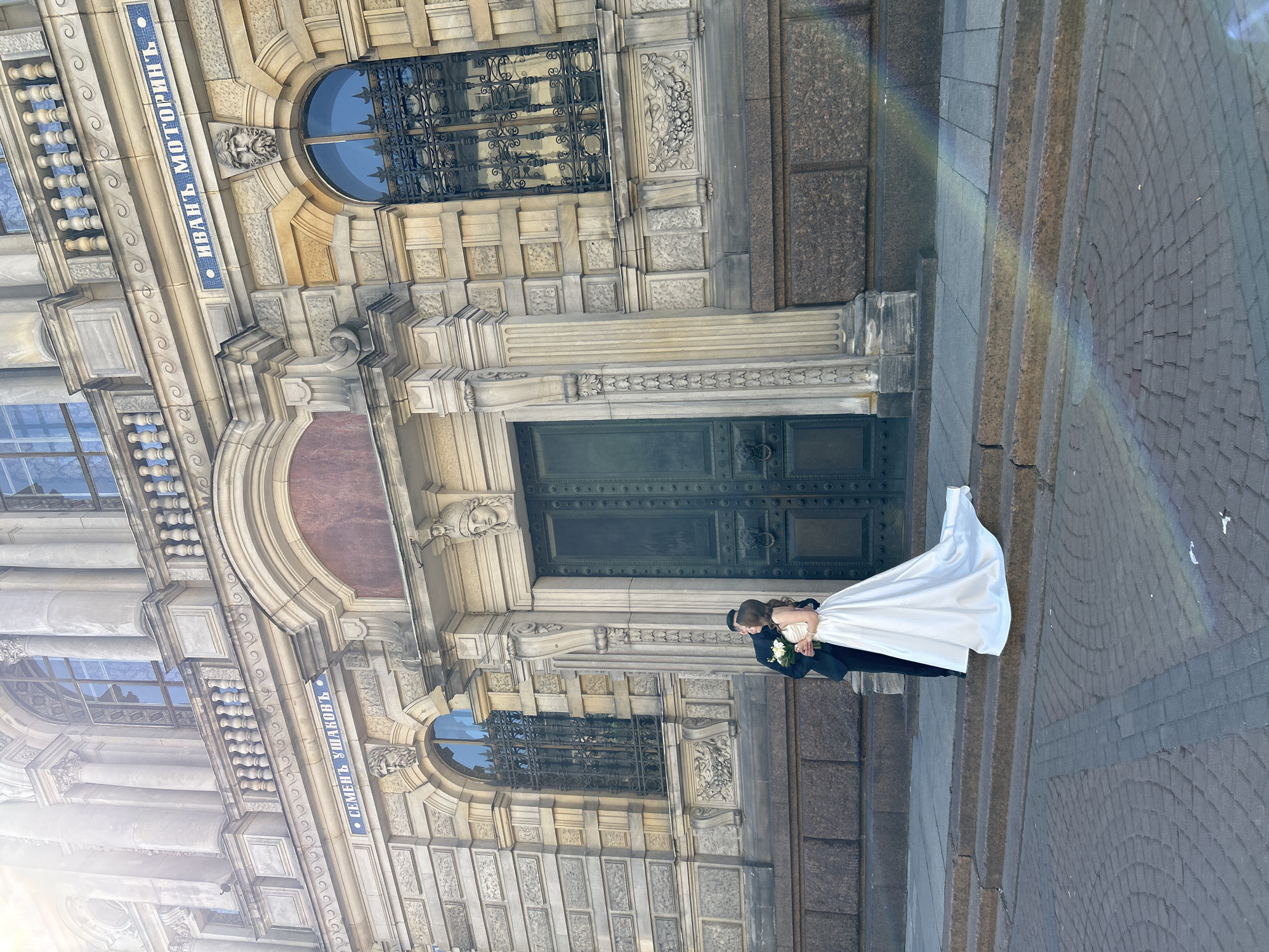 A couple is posing for a wedding photo in front of a building in Saint Petersburg, Russia. The bride is wearing a white dress and the groom is wearing a dark suit. They are standing on a cobblestone sidewalk in front of a large building with arched windows and a large dark door. There are signs in Russian on the building, one of which reads "Иванъ Моторинъ" (Ivan Motorin) and another, partially visible, "Семеиъ Ушаковъ" (Semey Ushakov).