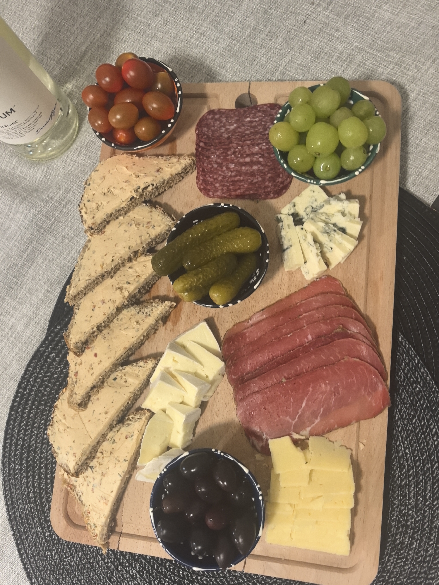 A high-angle, close-up shot shows a wooden charcuterie board with various foods arranged on it. Clockwise from the upper left, there is a small bowl of red and brown cherry tomatoes, sliced salami, a small bowl of green grapes, wedges of blue cheese, sliced ham, stacked slices of yellow cheese, a small bowl of black olives, wedges of white cheese, slices of bread with a spread, and a small bowl of pickles. A bottle of white wine is visible in the upper left corner of the image. The board rests on a grey woven placemat, and part of a light grey tablecloth is visible on the left side. The location is unknown and not within the scope of provided information.