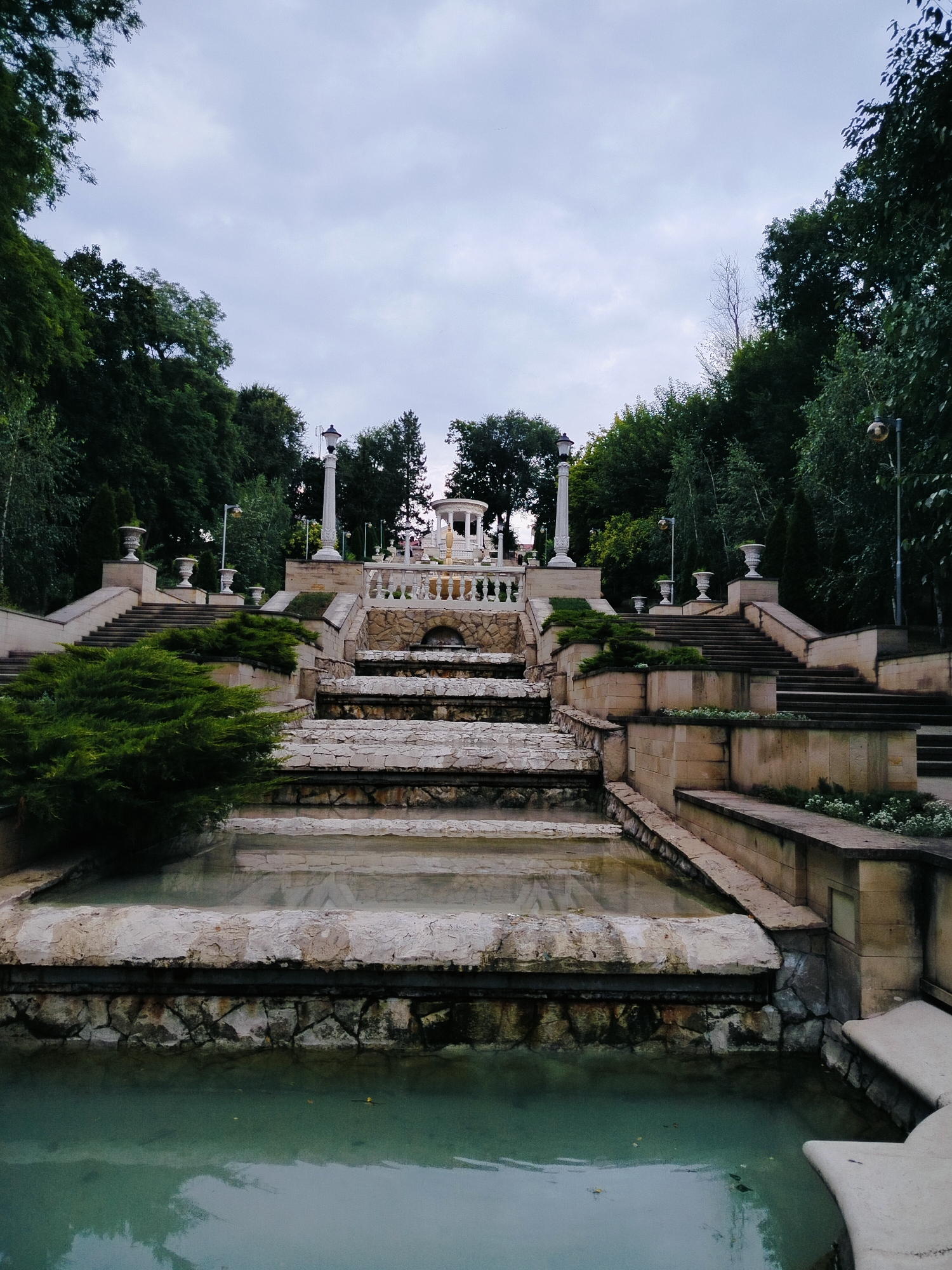 The image depicts the Cascade Staircase, a monumental architectural ensemble in Chișinău, Moldova. The scene is dominated by a series of tiered stone steps and water features, creating a terraced effect. Lush green trees and dense foliage frame the staircase on both sides, suggesting a park-like setting. The sky is overcast, contributing to a muted, somewhat somber atmosphere.
At the top of the staircase, a classical gazebo or rotunda is visible, adorned with white balustrades. flanking the upper portion of the stairs are tall, ornate lampposts and large, urn-shaped decorative vases. The stone construction of the staircase is evident, with a mix of rough-hewn stone and smoother, lighter colored elements. Small pools of water are integrated into the lower tiers, reflecting the cloudy sky and surrounding greenery. In the foreground, a larger pool of greenish-tinted water occupies the bottom of the frame. No people are visible in the image. The overall impression is one of grandeur and serene, albeit slightly melancholic, beauty.