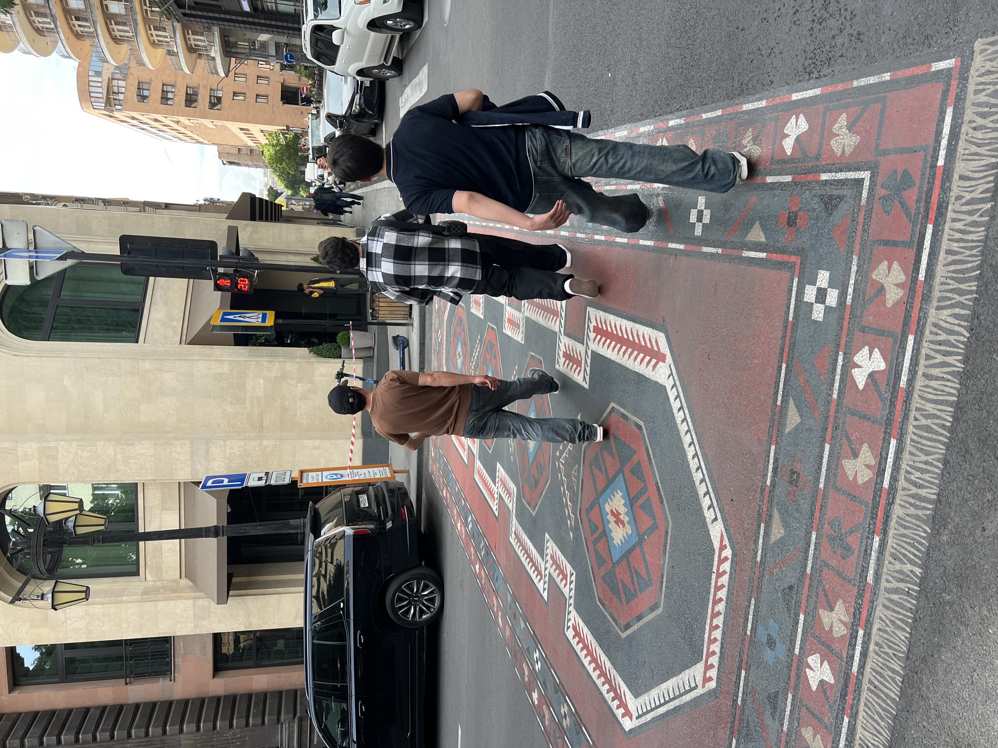 The image shows a street scene in Yerevan, Armenia, featuring a large decorative rug painted on the asphalt, serving as a crosswalk. Three men are walking across the rug from right to left. The first man is wearing a dark blue shirt and jeans, the second a black and white checkered shirt, and the third a brown shirt and jeans. Buildings line the street on both sides, with cars parked along the curb. A traffic light is visible on the left side of the street, along with street signs. The image is taken at street level.