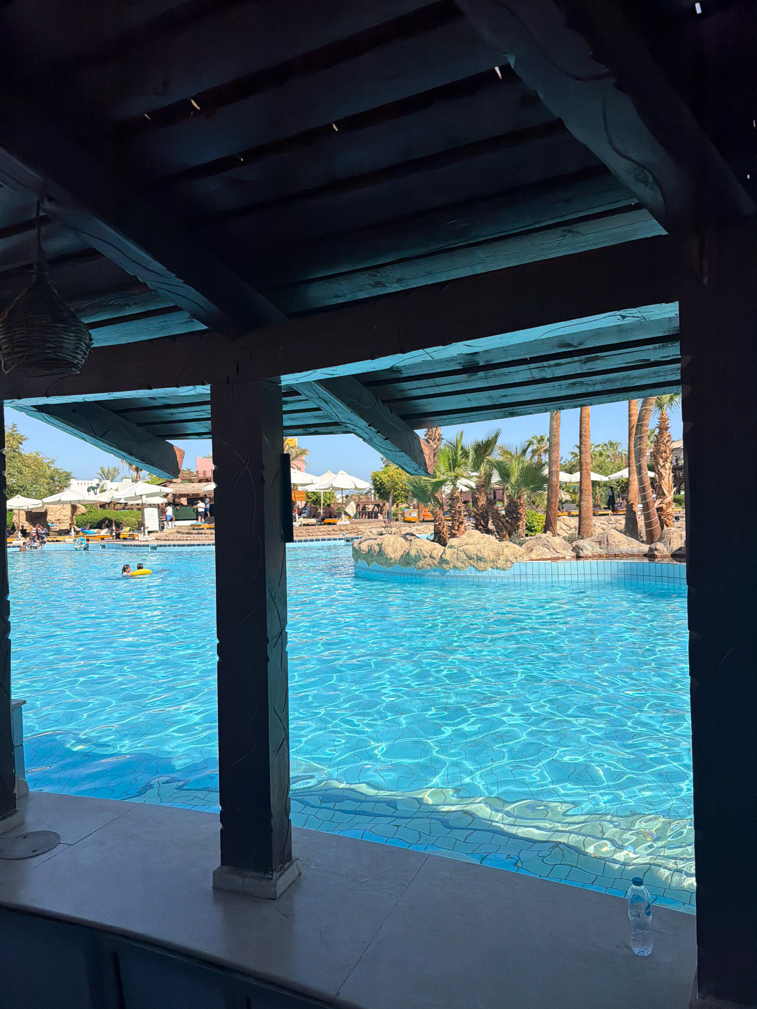 The image captures a sunny day at a resort pool in Sharm El Sheikh, Egypt. The foreground features a dark wooden structure, likely a bar or shaded seating area, with visible beams and a hanging woven basket light fixture. The view looks out onto a large, vibrant blue swimming pool, its surface shimmering with light reflections. In the pool, two individuals are visible: one person appears to be swimming, and another is sitting in a yellow inflatable ring.
Beyond the pool, the resort landscape unfolds. Numerous white umbrellas are open, providing shade for lounge chairs arranged on a tiled surface. Palm trees and other tropical foliage dot the area, adding to the resort ambiance. In the background, several low-rise buildings with terracotta-colored roofs are visible, interspersed with more vegetation. People are scattered throughout the scene, relaxing and enjoying the pool area. The sky is clear and bright blue, indicating good weather. A plastic water bottle is placed on the ledge in the foreground.