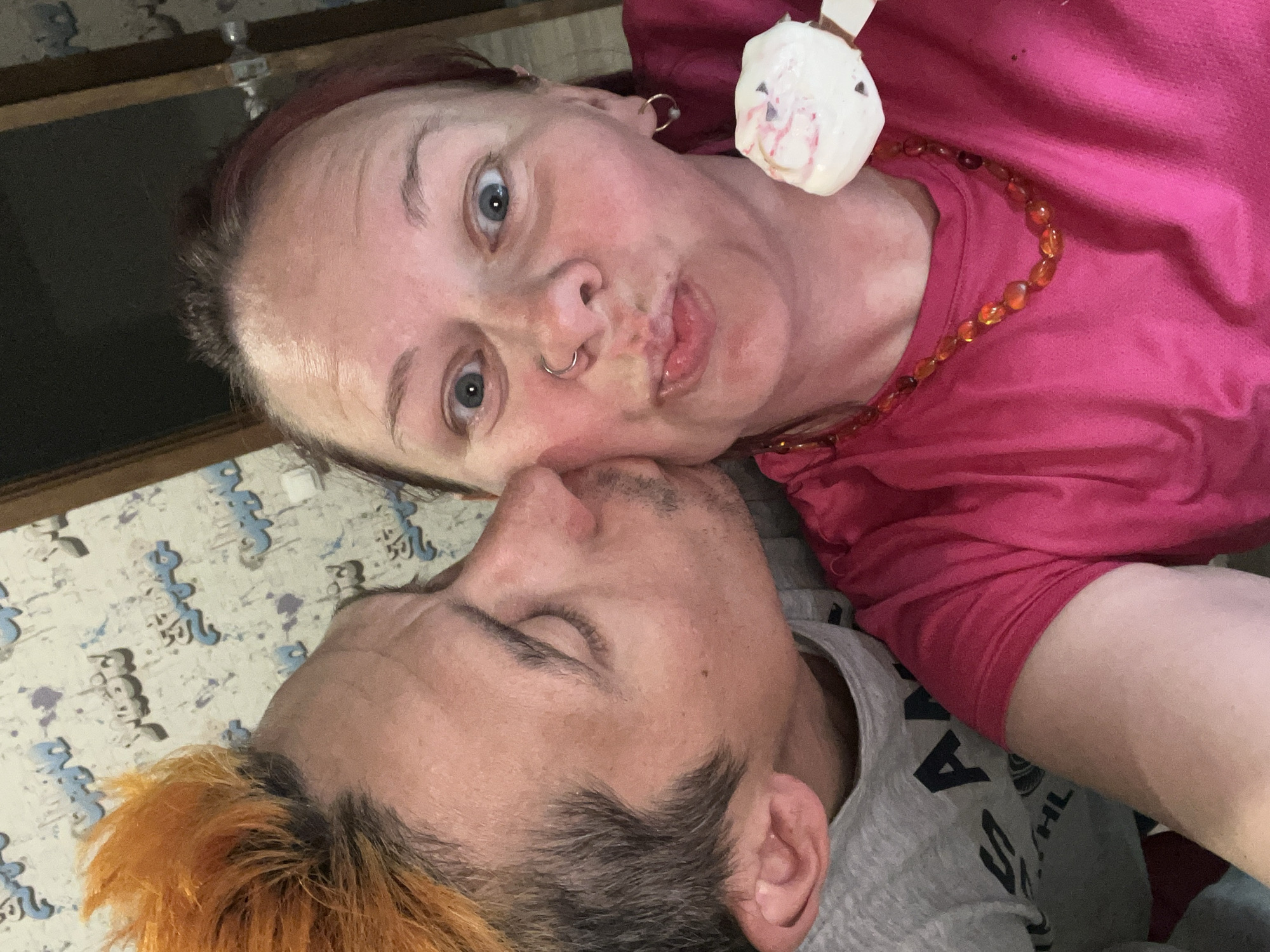 A selfie taken at a close angle shows a woman and a man lying on a surface with a light-colored patterned covering. The woman, wearing a pink top and an amber-colored beaded necklace, has fair skin, blue eyes, and a nose ring. She is making a pouting face towards the camera. A small, white object, possibly a toy or decoration, is positioned near her neck. The man, with orange-tipped hair, lies close to her with his eyes closed. He is wearing a gray t-shirt with partially visible text. The background is slightly out of focus. The location is Chaikovsky, Russia.