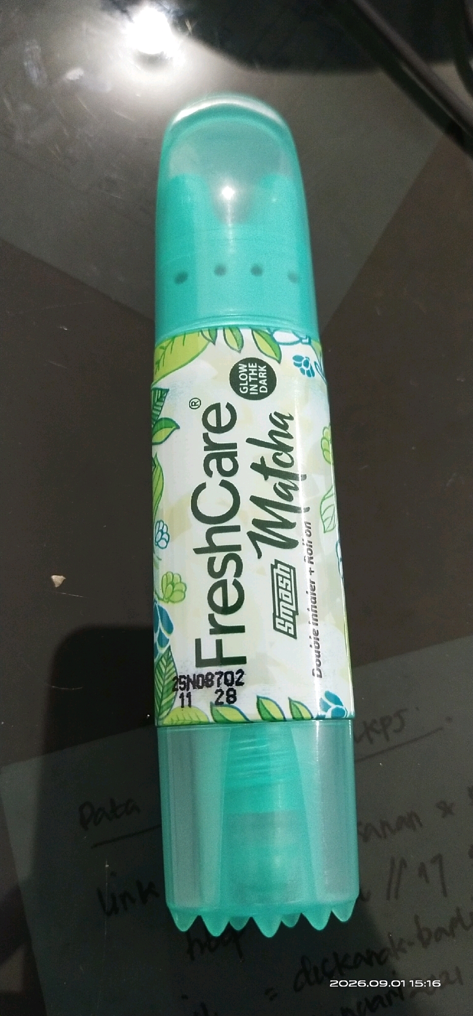 The image shows a close-up, eye-level, vertical shot of a teal-colored roll-on product with a patterned label featuring green leaves and flowers. The label text reads "FreshCare Matcha" and includes a small circular icon labeled "GLOW IN THE DARK". Below this, there is text that appears to say "Smash Double Immunity + Roll-on". A batch code and date stamp, "25N08702 11 28", are visible on the lower part of the label. The product is resting on a dark, reflective surface, possibly a table. In the background, out of focus, are handwritten notes on paper, one of which appears to be a date stamp "2026.09.01 15:16". The lighting suggests an indoor environment. The image does not contain any information or visual cues that would allow for the determination of its location in Tamiang Layang, Indonesia.