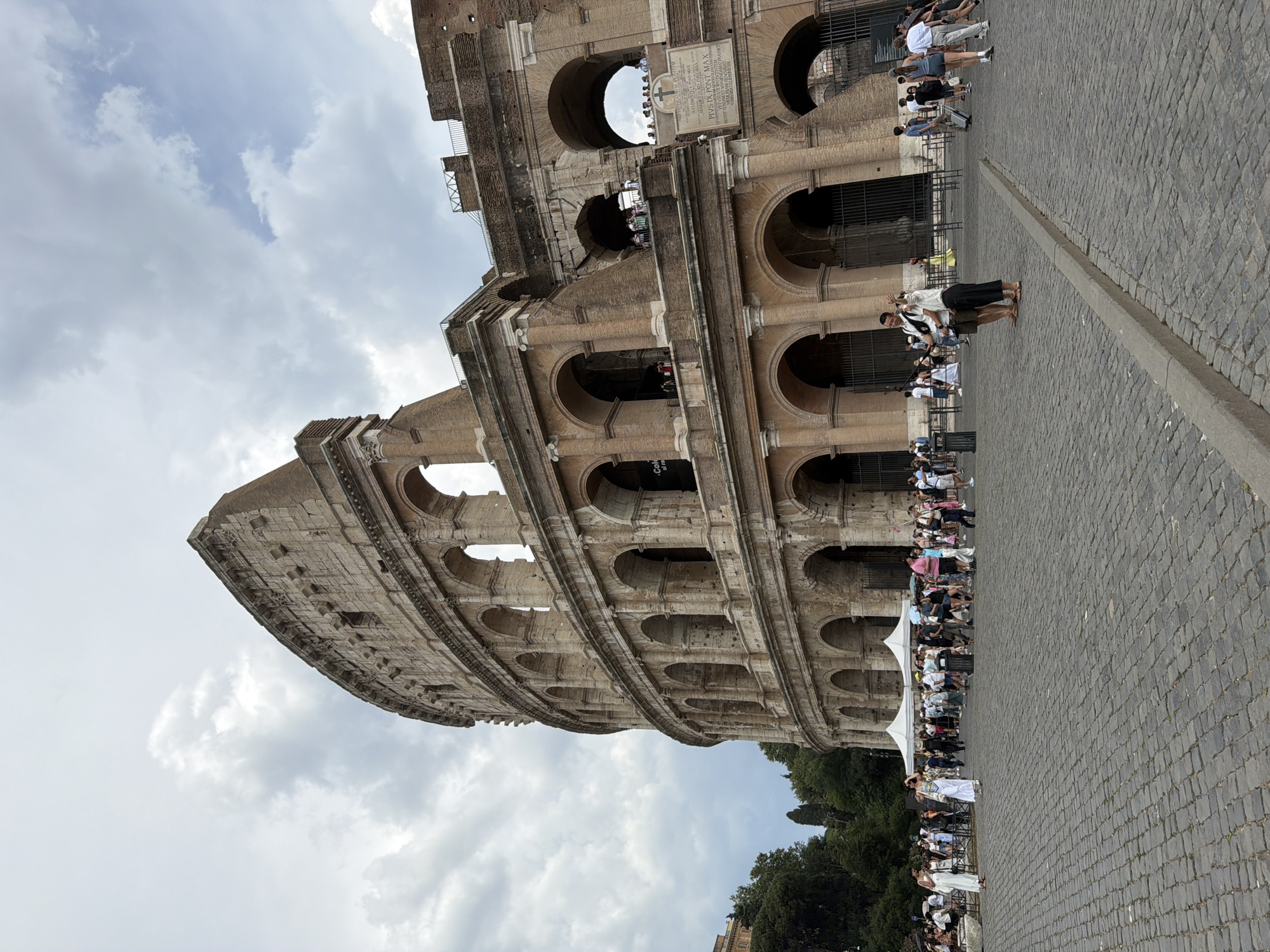 This is a vertical, eye-level, medium shot of the Colosseum in Rome, Italy. The sun is out, but there are a lot of clouds, suggesting a partly cloudy day, likely late morning or early afternoon. The Colosseum itself takes up a large portion of the frame, with its iconic arches and weathered stone structure. In the foreground, a crowd of tourists is visible, with some people standing and others seated. They are gathered on the cobblestone pavement in front of the monument, with some individuals appearing to take photos or observe the architecture. A few white tents are set up, possibly for shade or vendors. The text "Col" and other inscriptions are visible on a plaque attached to the Colosseum's structure. The overall scene captures a typical tourist experience at a historical landmark.