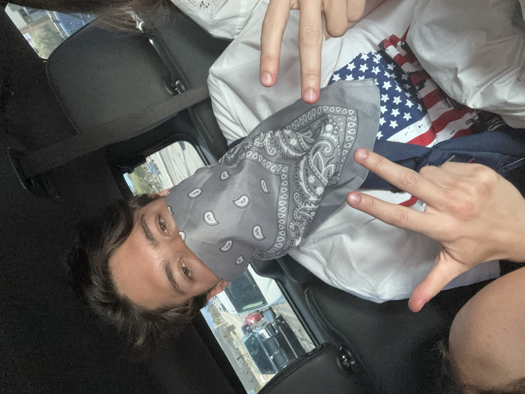 A man is in a car wearing a gray paisley patterned bandana over his nose and mouth. His light brown hair is long, and his eyes are brown. He is wearing a white shirt with an American flag bandana beneath it. His hands are in the peace sign position. The car is black, and there are other cars visible through the window, as well as a white building. The location is unknown.