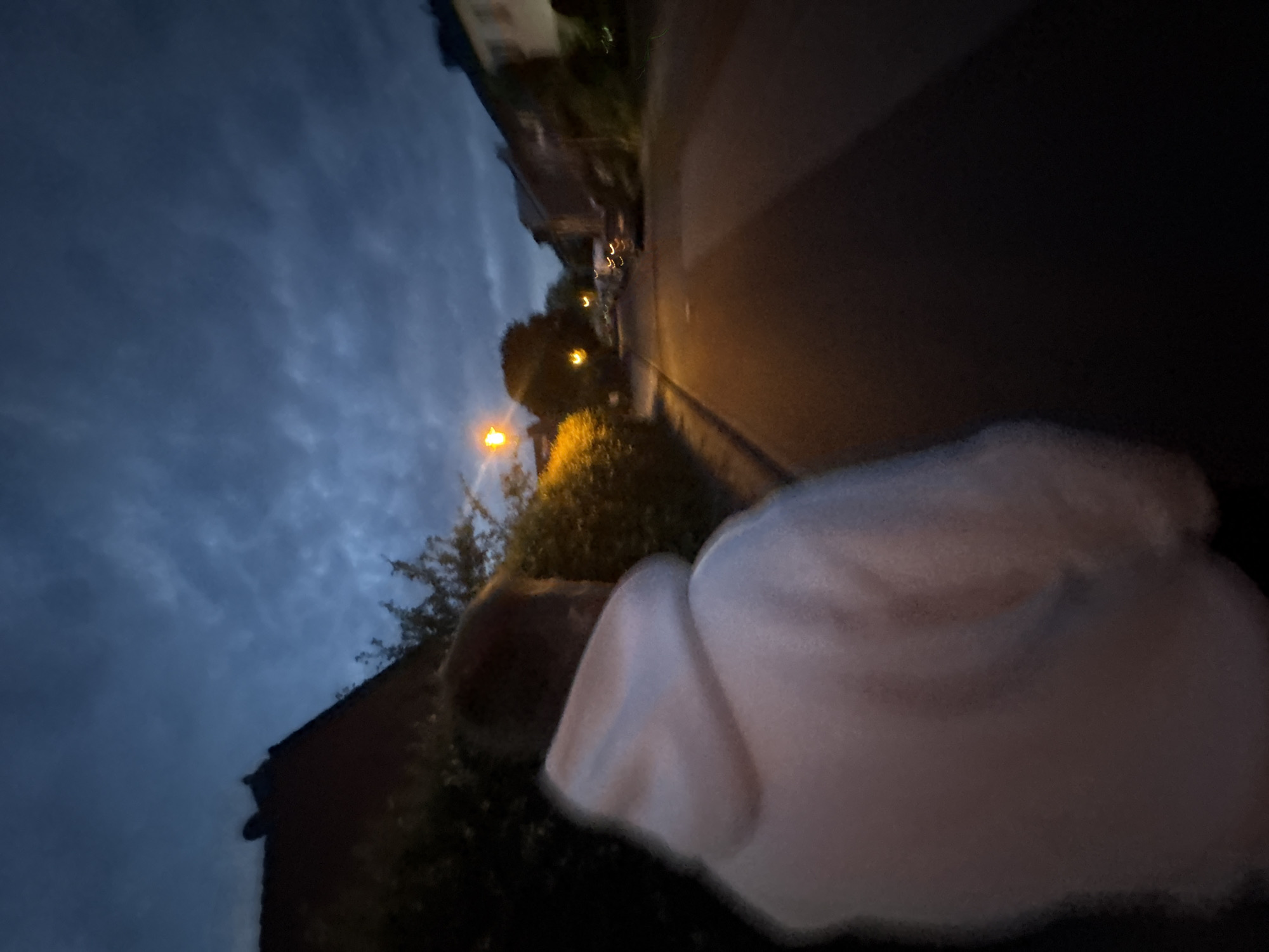 The image shows a person from behind, wearing a white hooded sweatshirt, walking on a street at dusk. The street is lined with houses and trees. A street lamp is illuminated, casting a warm glow on the surroundings. The sky is dark with clouds, suggesting an approaching night. The scene appears to be a residential area in Mönchengladbach, Germany, possibly a quiet street with houses on either side. The person is the sole human subject in the frame, and their face is not visible. The overall atmosphere is calm and somewhat subdued, typical of an evening walk.