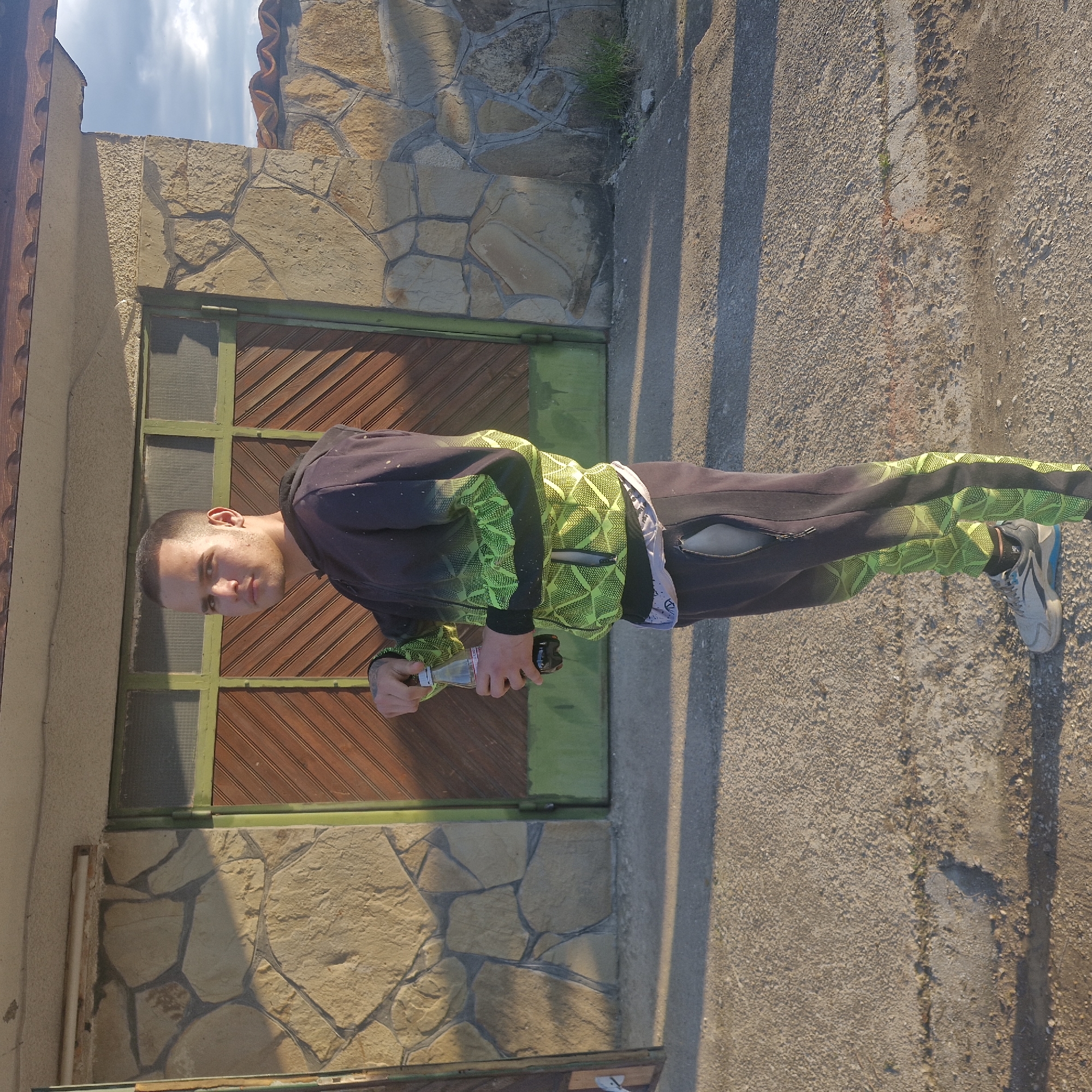 A man in a black and green tracksuit stands outside a stone building with a brown wooden door in Hirevo, Bulgaria. He is holding a bottle. The area is paved. The building is a single story. The sky is visible.