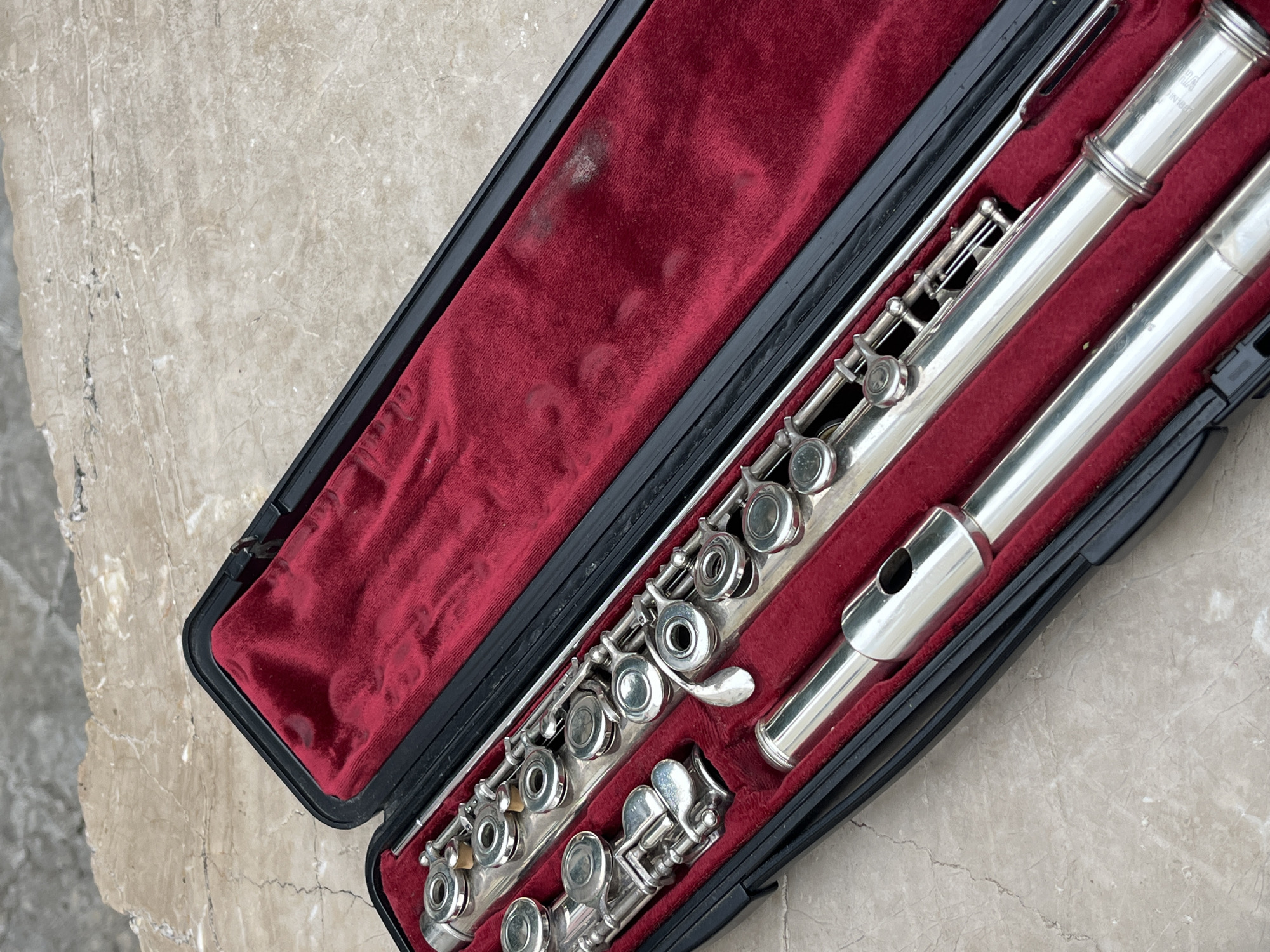 A silver flute is nestled in a black case with a red velvet interior. The case is open and rests on a light beige surface with subtle veining. The background and the lack of specific location markers prevent definitive determination of the location of the city of Budva, Montenegro.