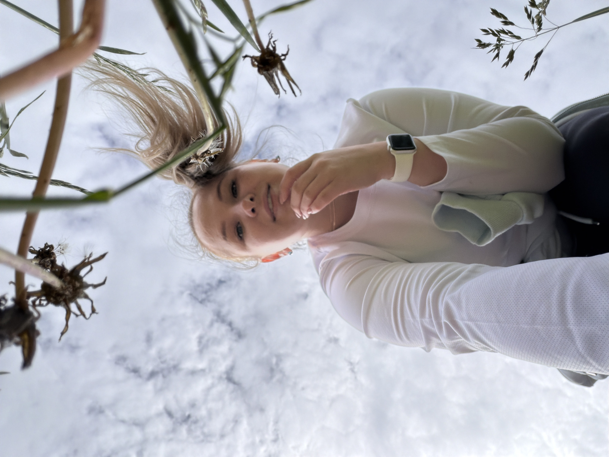 A woman with fair skin and blonde hair pulled back in a ponytail poses for a photo from a low angle, with her face and upper body in the frame against a backdrop of a cloudy sky. She is wearing a white long-sleeved shirt and a white smartwatch on her left wrist. Her right hand is near her face. Blades of grass and dead flower heads are scattered around the edges of the frame. It appears to be an outdoor setting. The location is Ольховка, Russia.