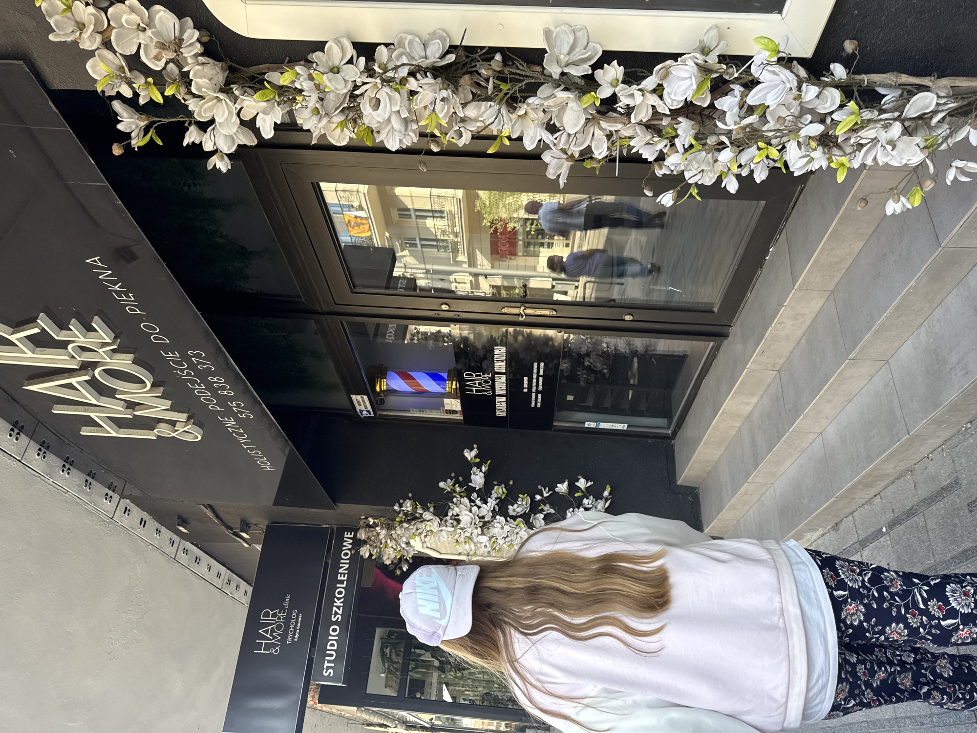The image shows the exterior of a business in Poznan, Poland, likely a salon or cosmetic clinic. The storefront features a black facade with illuminated signage that reads "HAIR & MORE" in large white letters, with smaller text below in Polish, including "HOLISTYCZNE PODEJŚCIE DO PIĘKNA" (Holistic approach to beauty) and a phone number "575 838 373". There is also a smaller black sign to the left that says "STUDIO SZKOLENIOWE" (Training Studio). A woman with long blonde hair, wearing a light-colored hoodie and patterned pants, is walking away from the camera towards the entrance. She is wearing a light blue Nike cap. The entrance has double glass doors with reflections of a building and people on the street. A barber pole with red, white, and blue stripes is visible inside the entrance on the right. Decorative white flowers with green leaves are attached to the right side of the facade, extending upwards. The ground in front of the entrance consists of concrete steps and a sidewalk. In the reflection of the door, two individuals are seen walking away from the building.