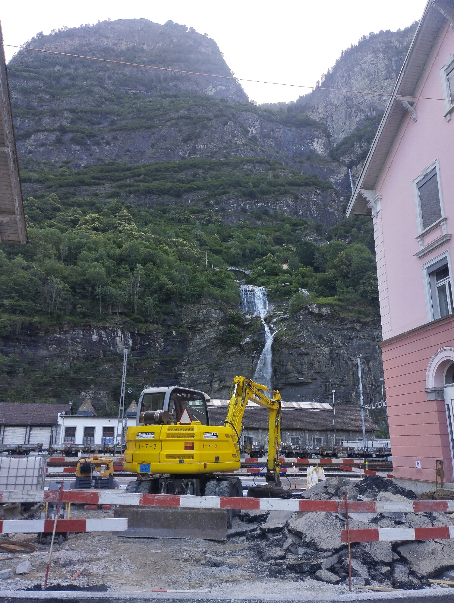 In an outdoor scene, a yellow excavator is positioned in a construction area in front of a pink building and a steep, forested mountain. The mountain features a waterfall cascading down its rocky face. The construction area is cordoned off with red and white striped barriers. Debris, including broken asphalt, is scattered across the ground. In the background, several buildings and train cars are visible. The sky is overcast.